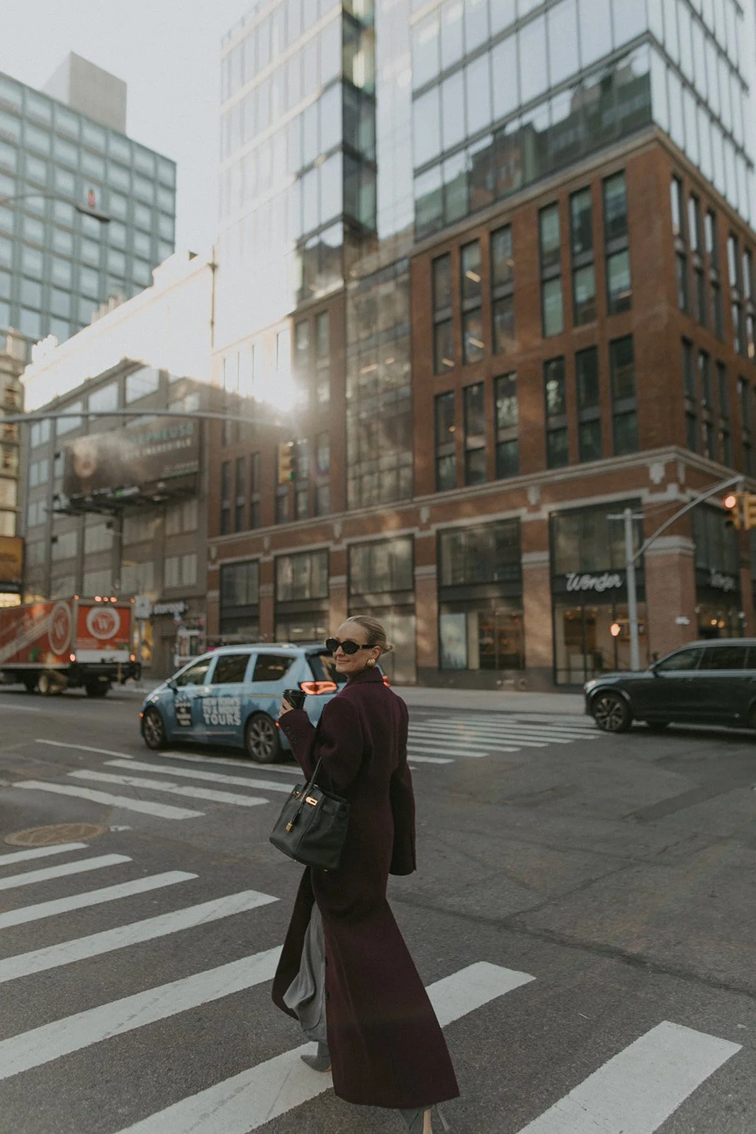 Virginia Bundy with sunglasses, wearing a long dark coat, crossing a New York City street at a crosswalk, with tall buildings in the background and cars passing by.