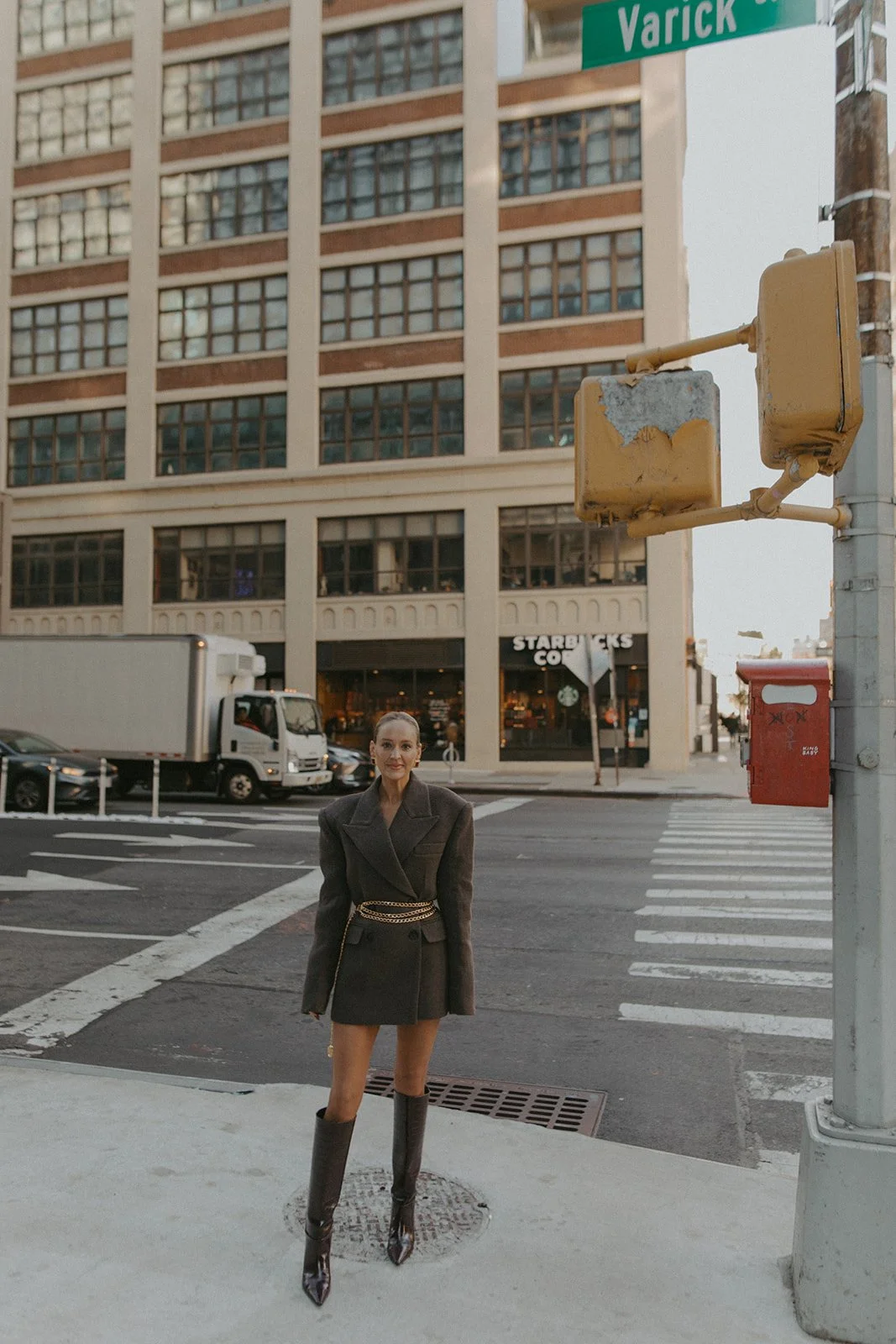 Virginia Bundy stands at a crosswalk in front of a large building with a Starbucks coffee shop on the ground floor in an urban setting. She is wearing a dark blazer dress with chain details and high-heeled boots, posing for the photo.