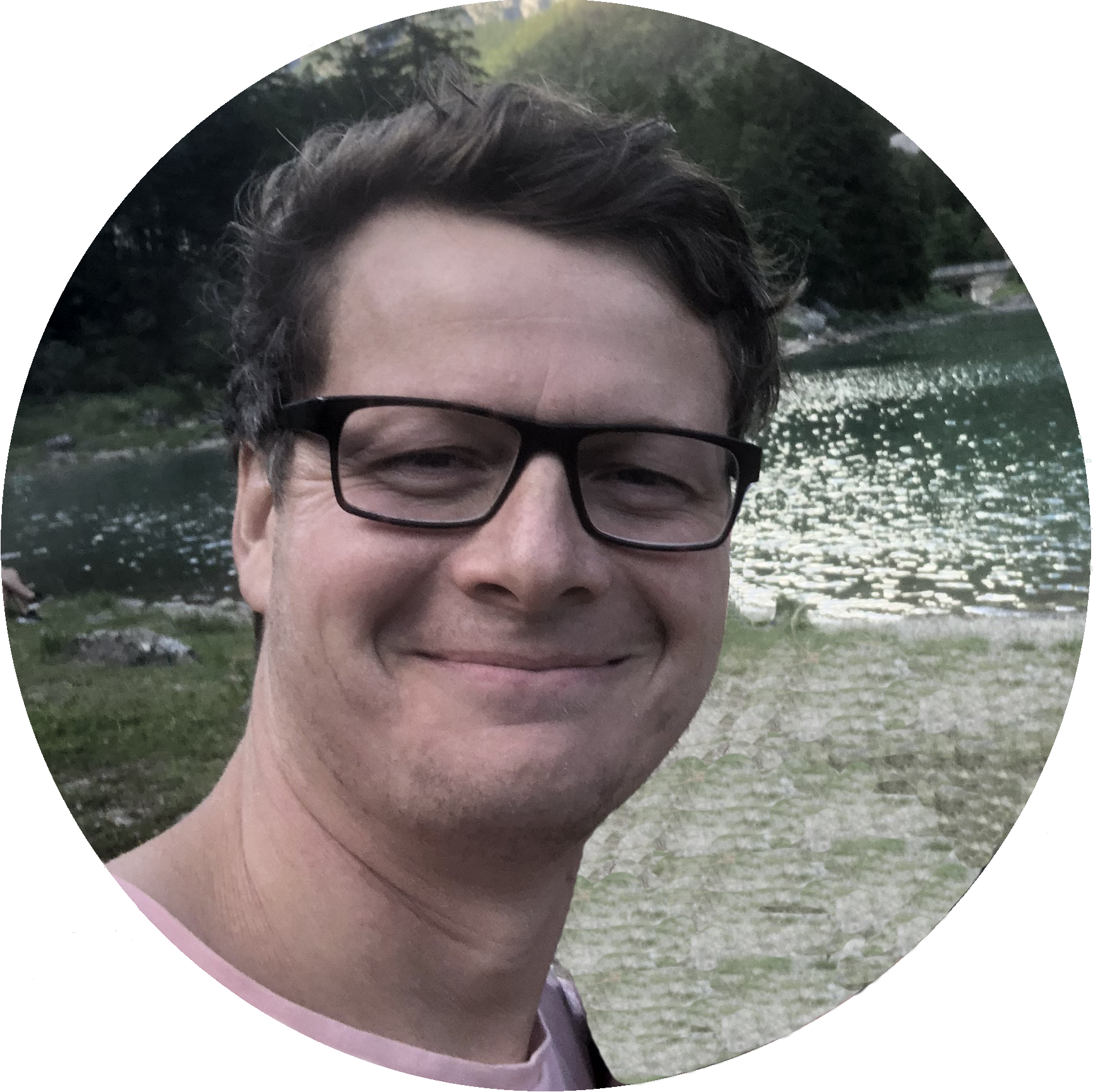 Nahaufnahme eines lachenden Mannes mit schwarzen Brillen an einem See mit Bäumen im Hintergrund.