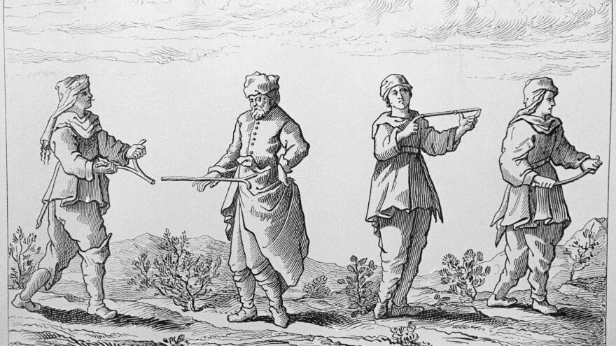 Vier Männer gehen in einer Reihe im Freien, einige halten Gegenstände in den Händen, die ihnen beim Gehen helfen, mit Bergen und Wolken im Hintergrund.