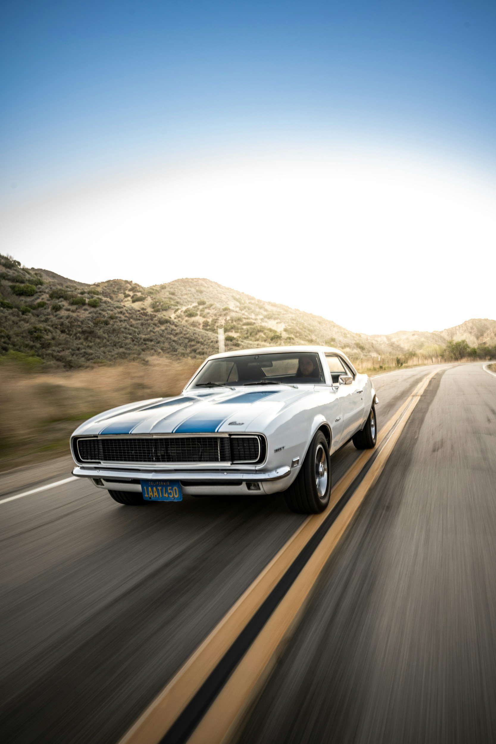 A white vintage car with blue racing stripes driving on a winding road in a desert landscape during daytime.
