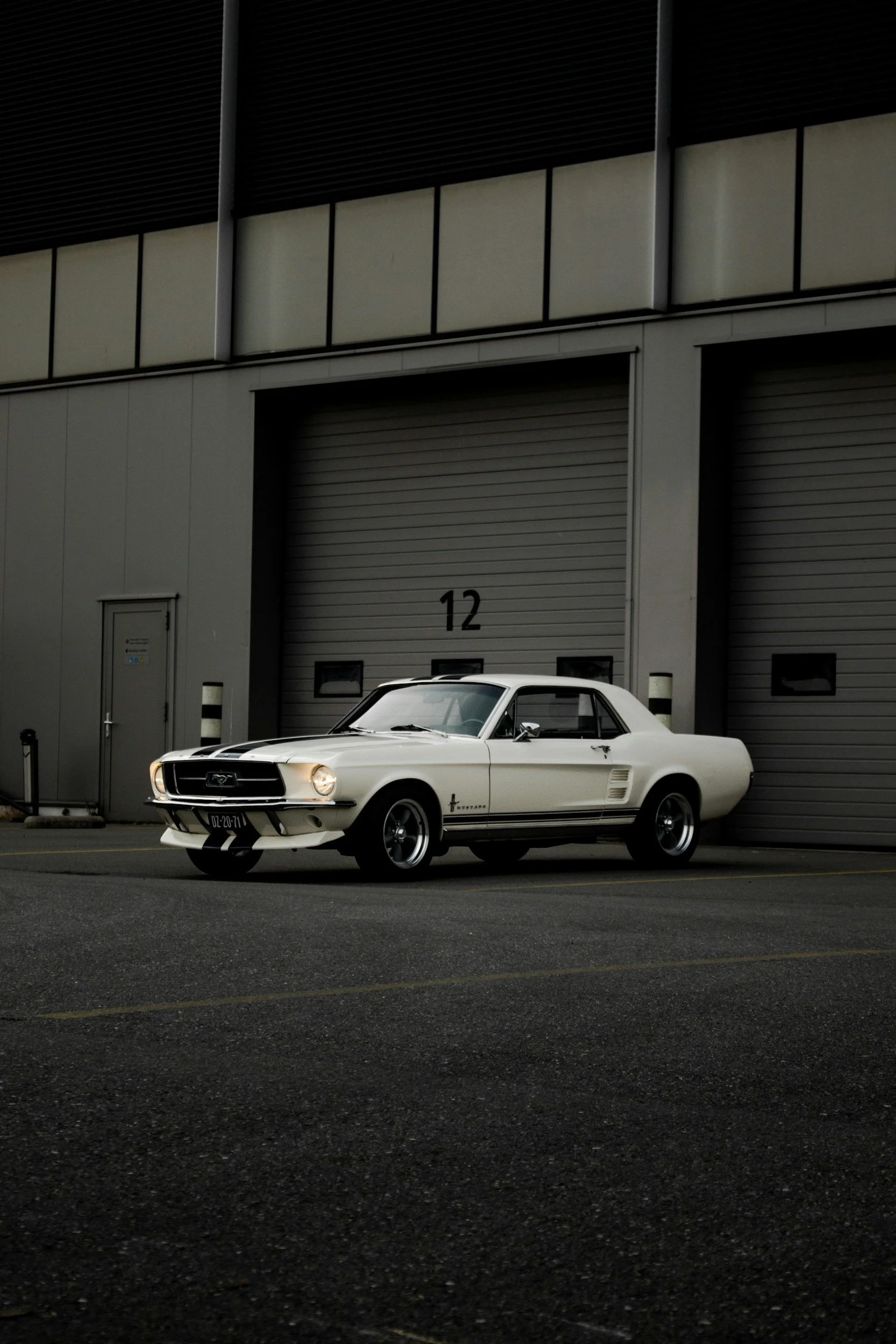 A vintage white Ford Mustang parked in front of a gray industrial building with large rolling doors and a number 12.