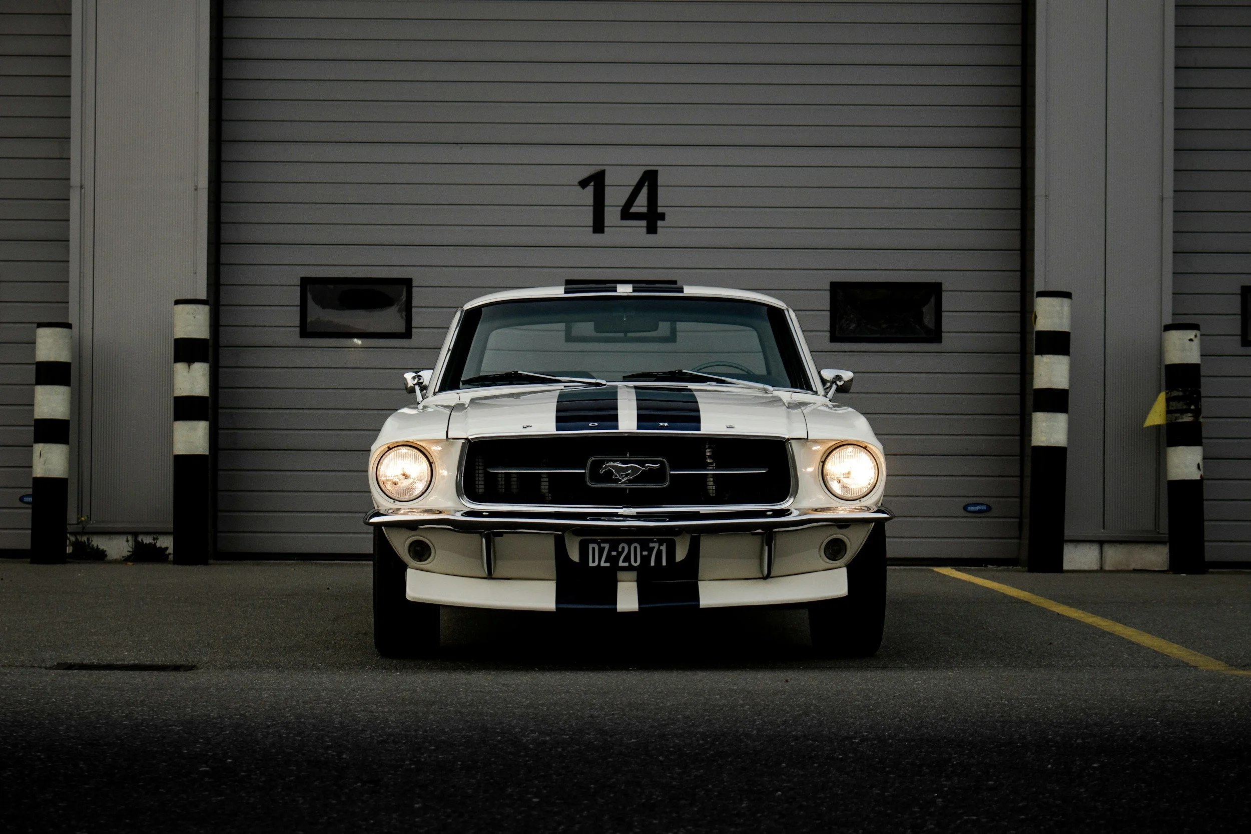 A white vintage Ford Mustang car with black racing stripes parked in front of a gray garage door marked with the number 14.