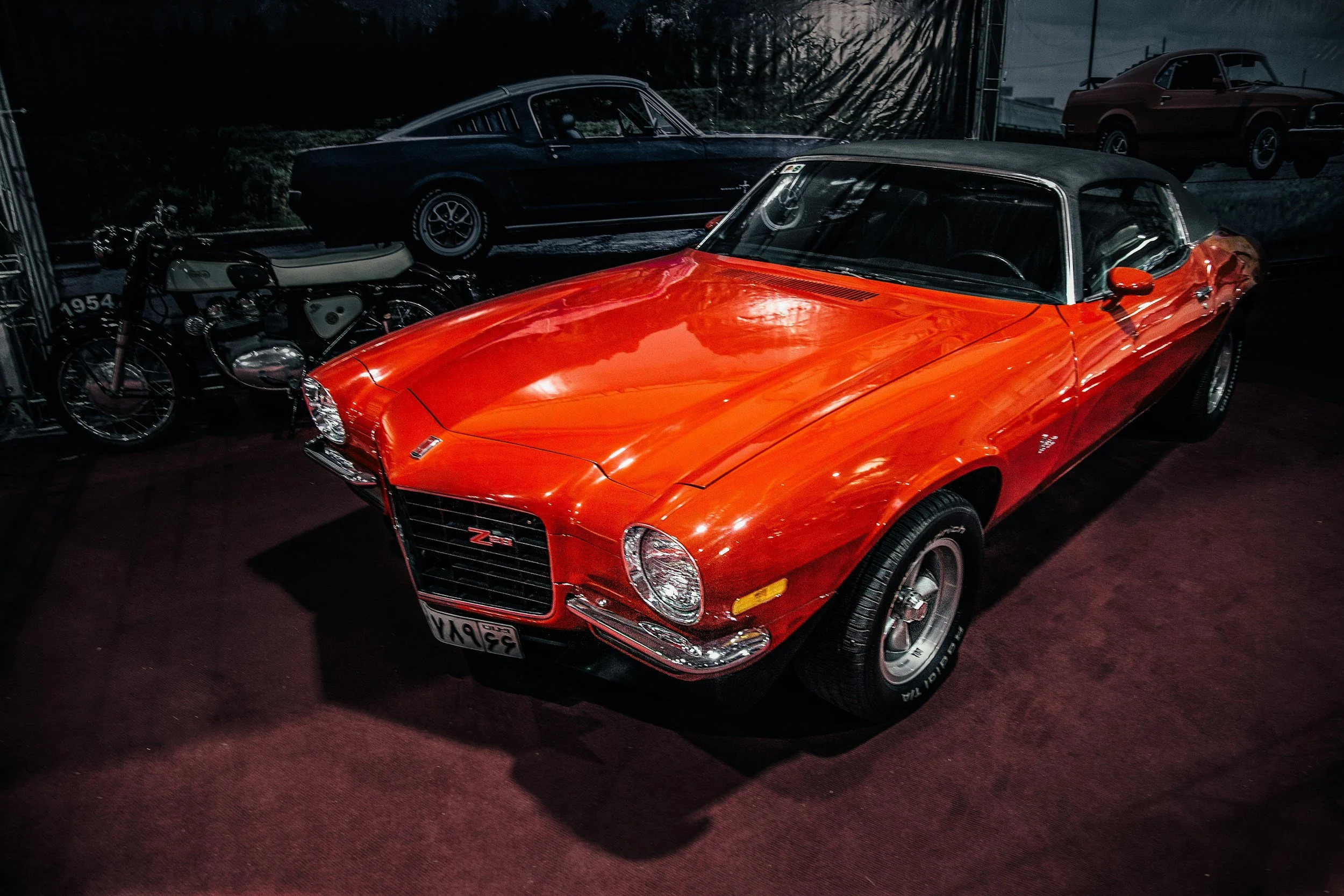 A bright orange vintage Chevrolet Camaro Z28 with a black convertible top on display indoors, with classic cars and motorcycle in the background.