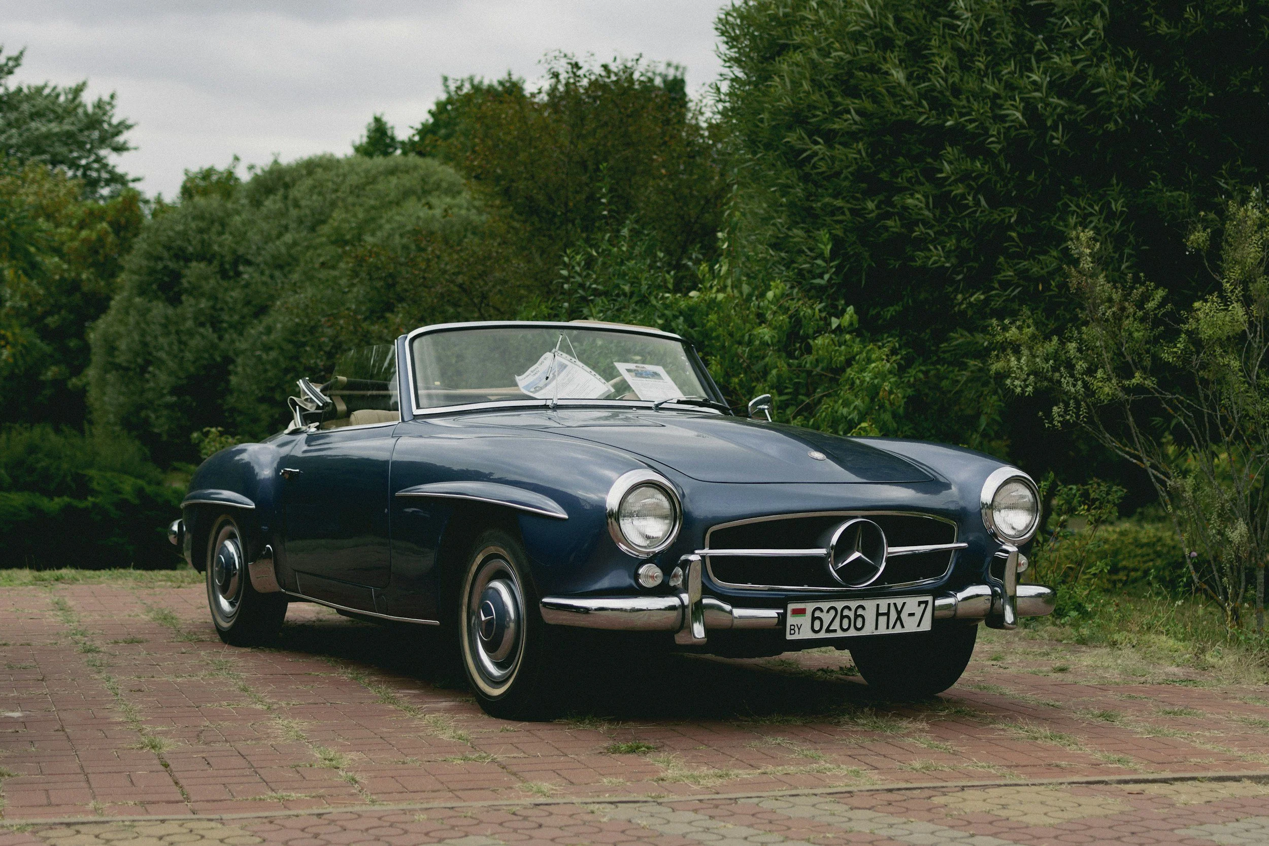 A vintage dark blue Mercedes-Benz convertible parked on a brick pavement, with green trees and bushes in the background.