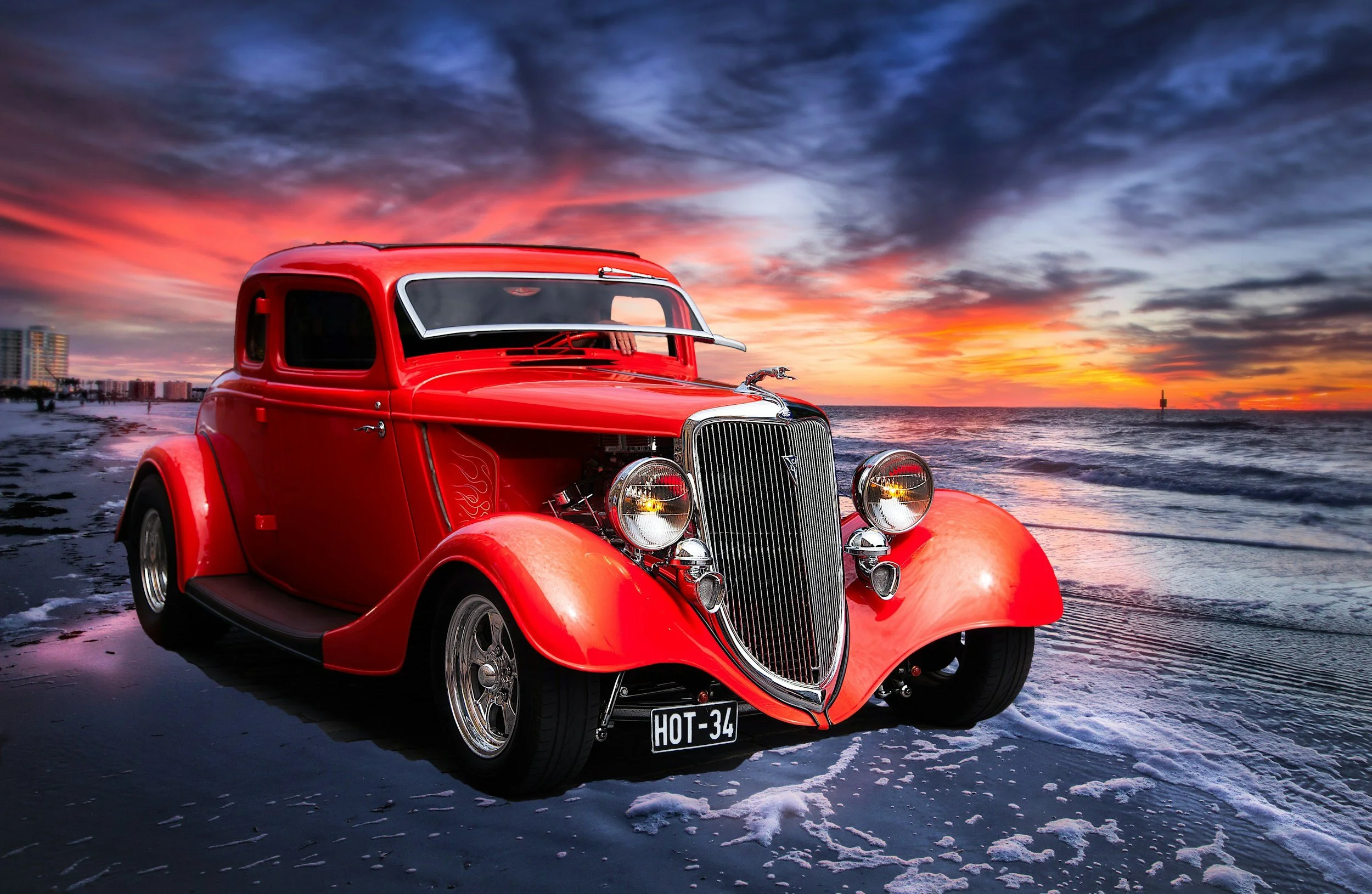 A vintage red car parked on a beach at sunset with colorful sky and ocean in the background.