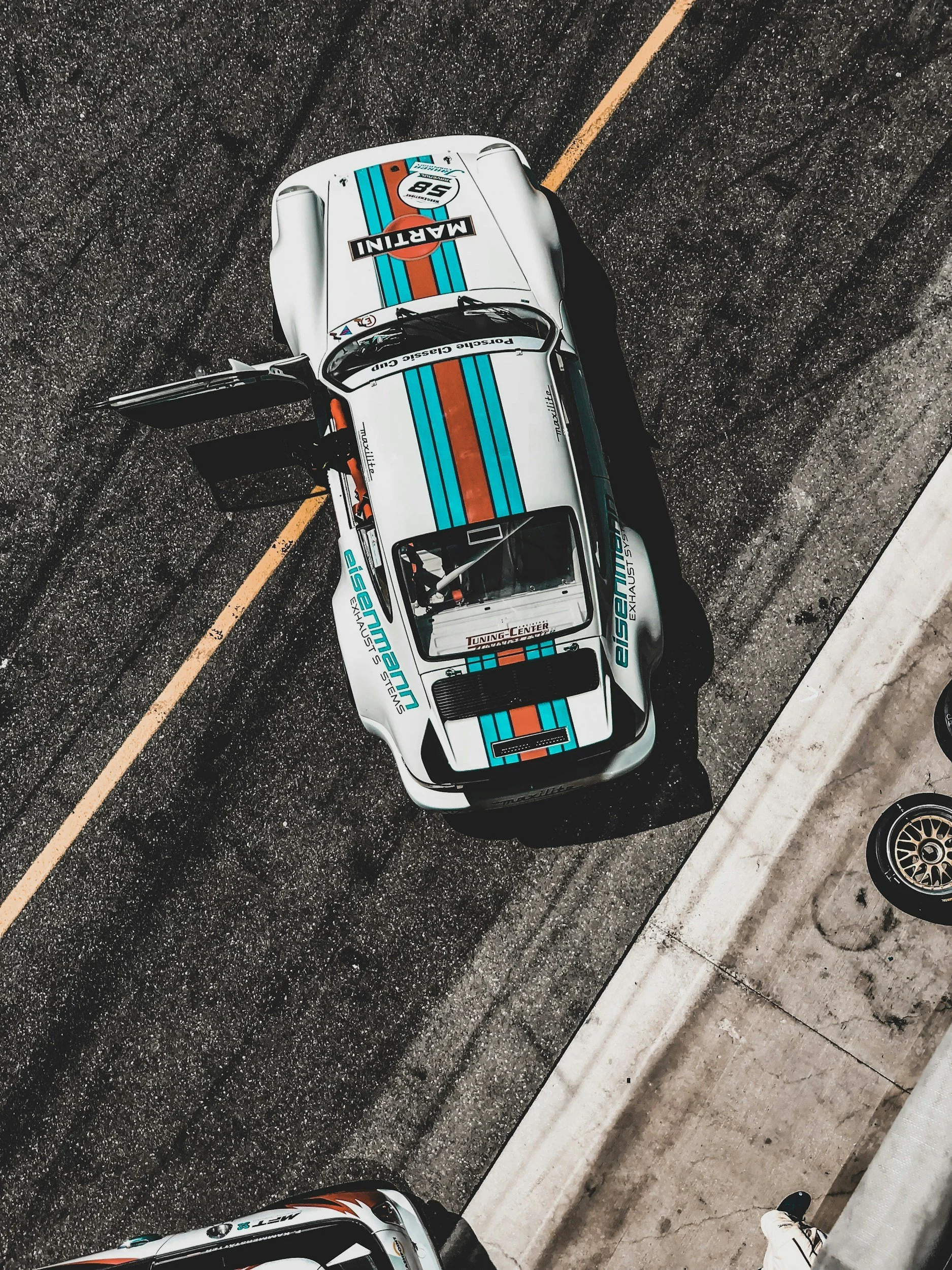 white race car with 2 doors blue and brown strips door open on racetrack picture from above
