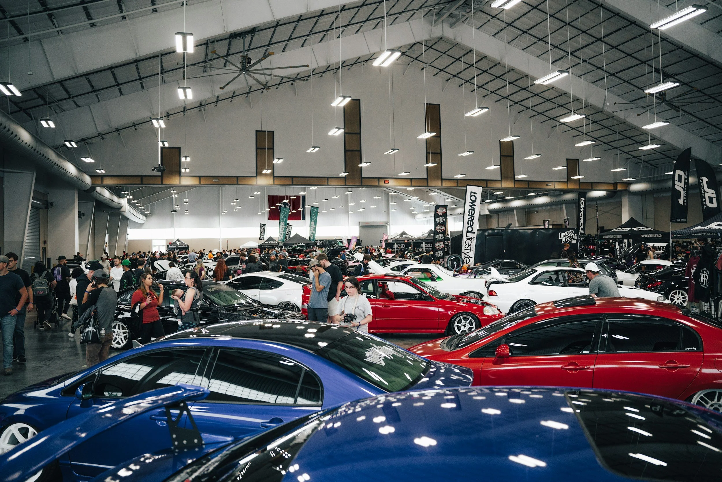 Interior of a large car show with various sports and luxury cars on display, and a crowd of people walking and talking.