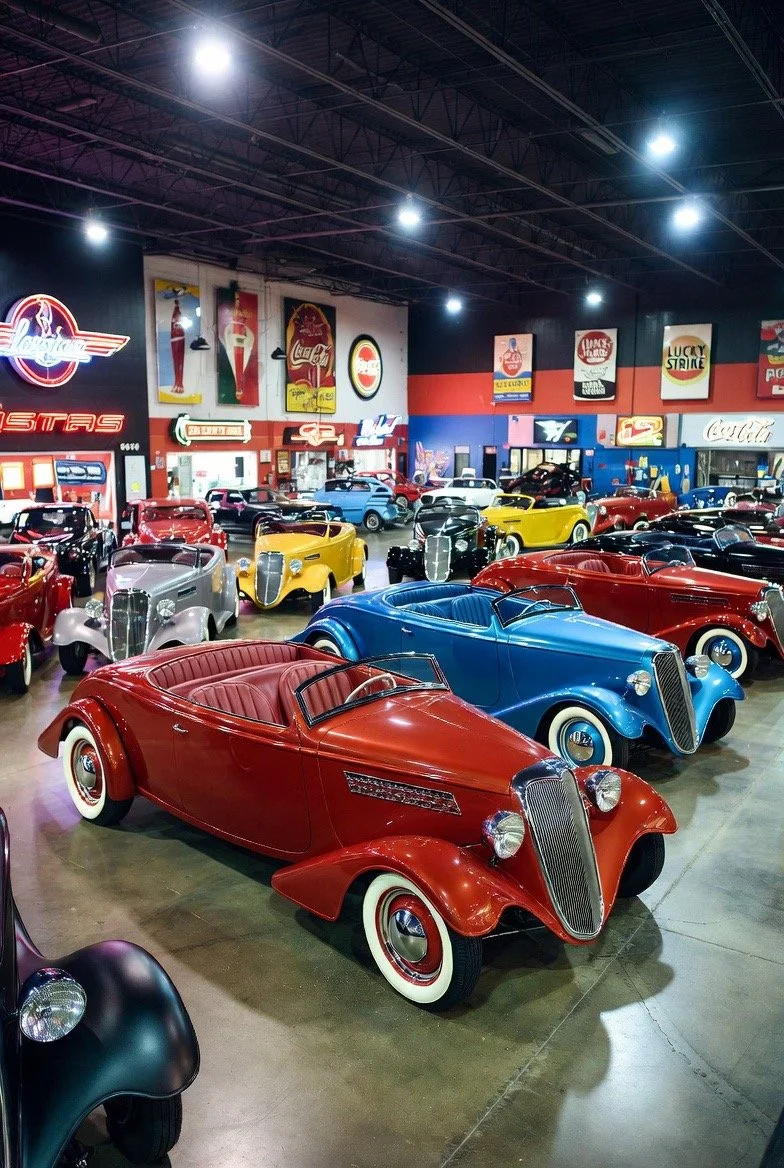 A vintage car showroom with several classic cars in red, blue, yellow, black, and gray, displayed indoors with neon signs and posters on the walls.