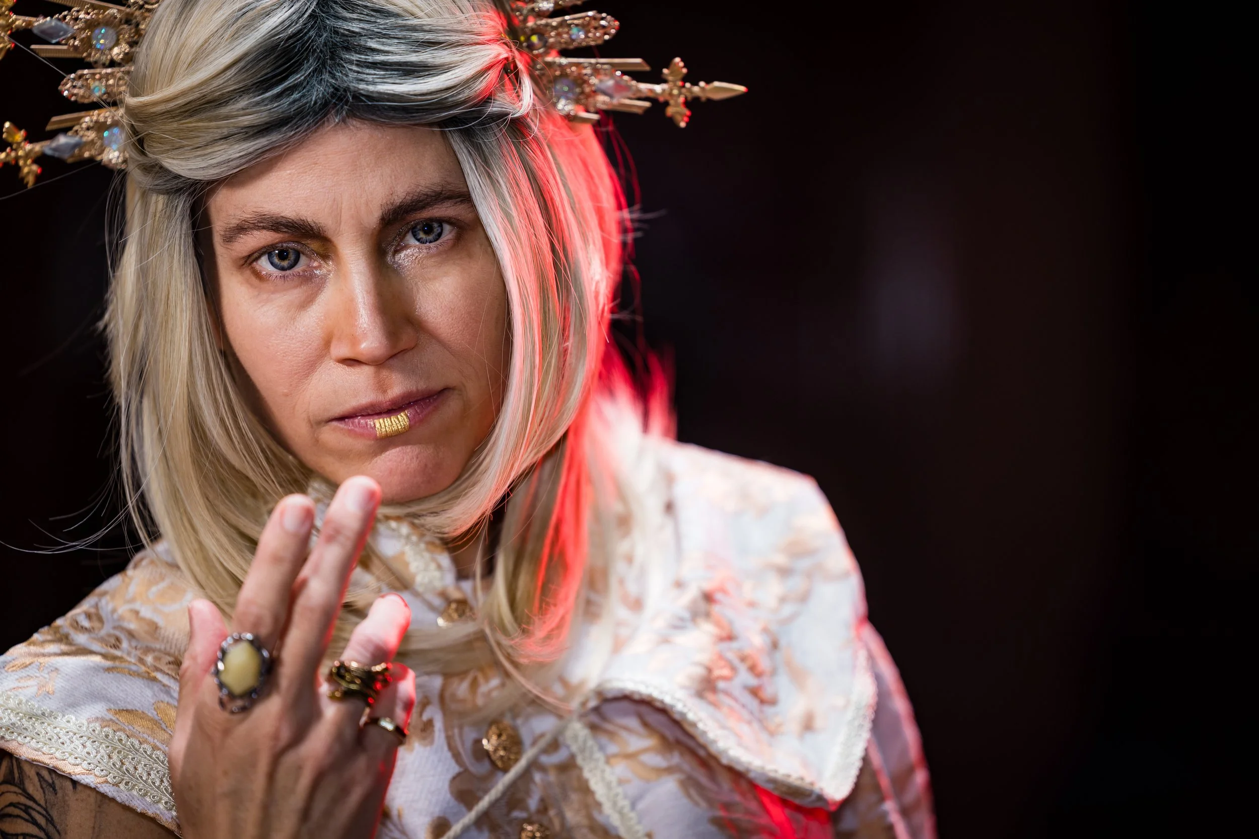 A woman with blonde hair wearing ornate clothing, multiple rings, and a gold lip ornament, holding up her hand with three fingers against a dark background with red lighting.