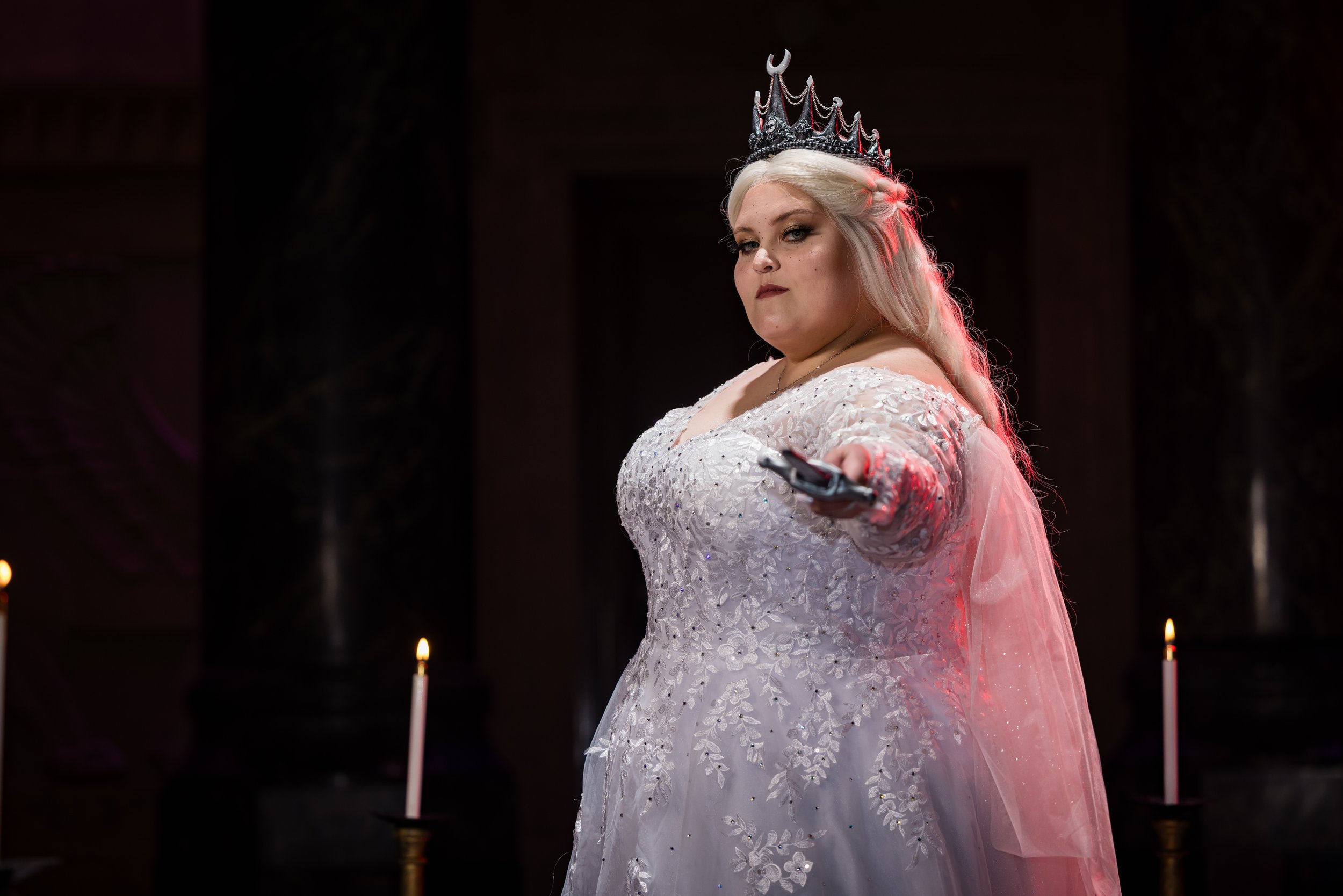 A woman dressed in a white, embroidered gown with a sheer cape, wearing a crown, and holding a gun pointed toward the camera, standing in a dark room with lit candles.
