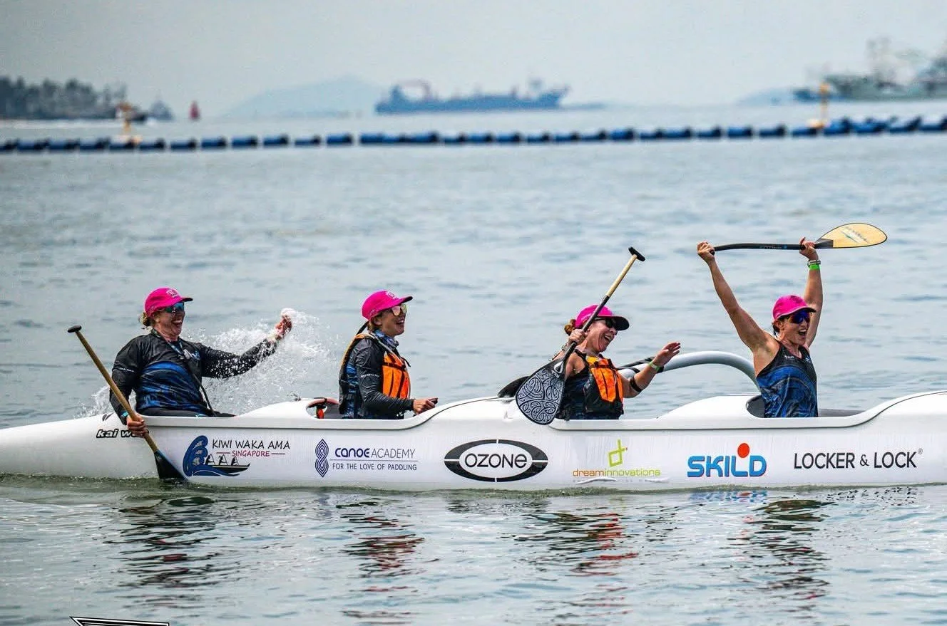 Four women in a white kayak, wearing pink hats and sunglasses, paddling in a body of water with a ship and distant shoreline in the background.