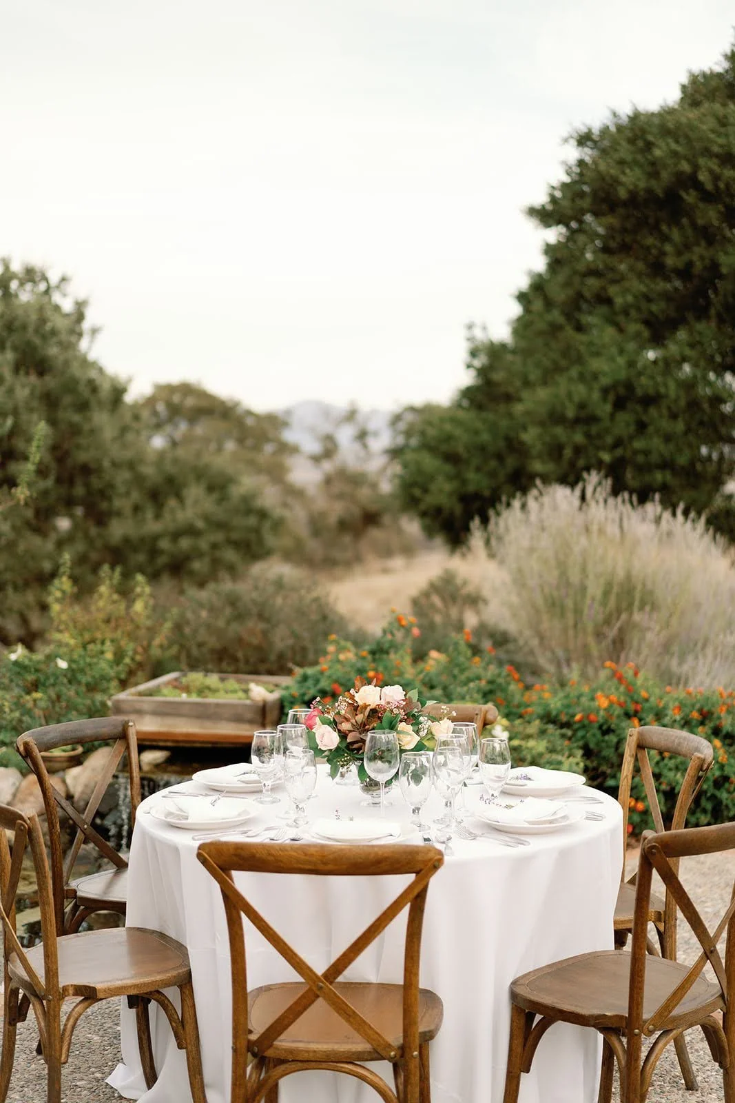 An outdoor dining setup with a round table covered in a white tablecloth, surrounded by six wooden chairs, and a floral centerpiece, set in a garden with trees and flowering bushes in the background.