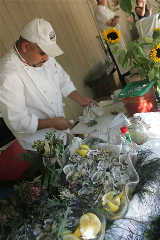 A chef in a white uniform and hat preparing fresh oysters on a table decorated with sunflowers and lemon slices, in a bright indoor setting.