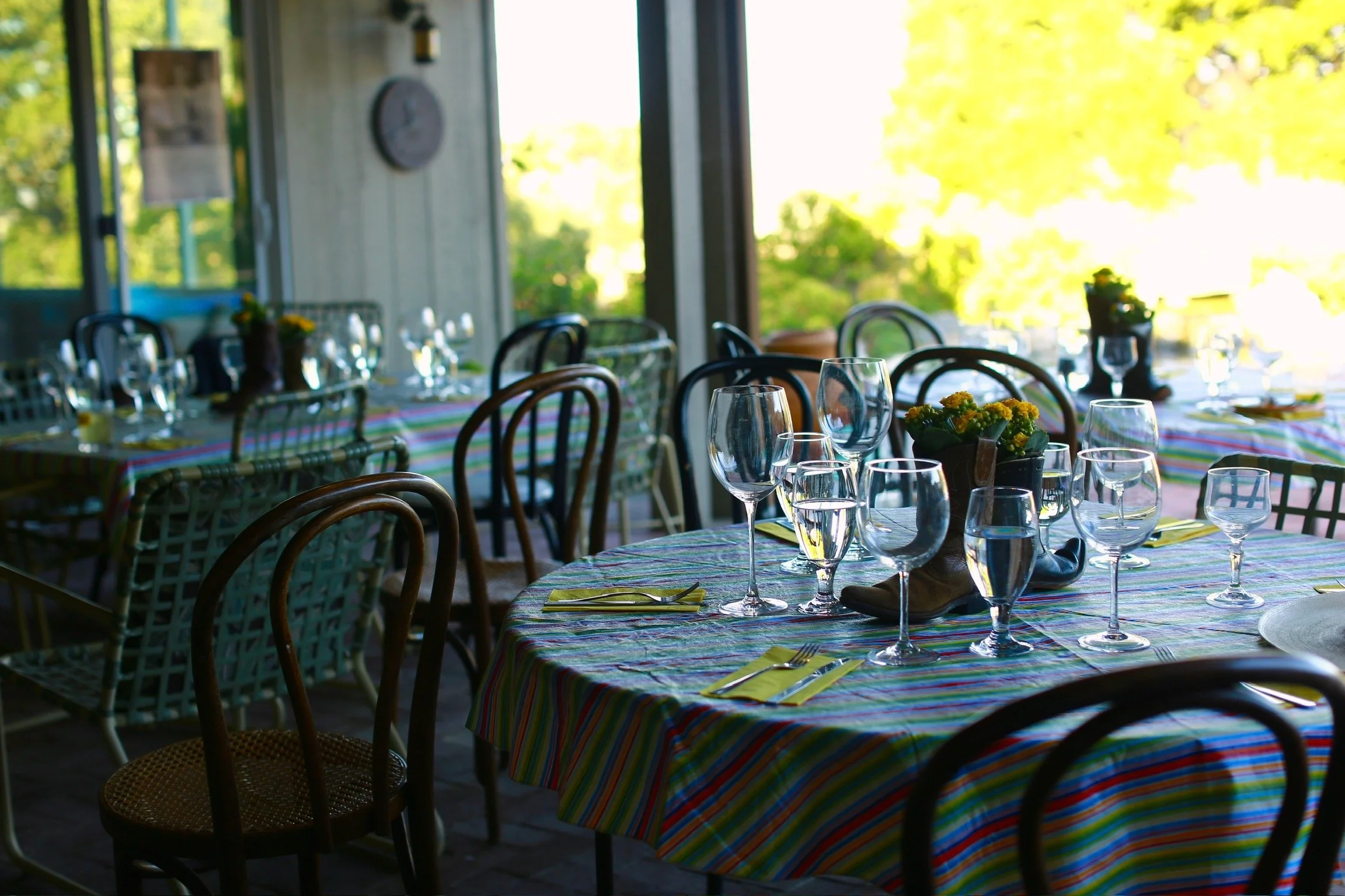 A dining area with tables covered in colorful, striped tablecloths. The tables are set with multiple wine glasses, water glasses, and yellow napkins with forks and knives. There are small floral centerpieces on some tables, and natural light floods i