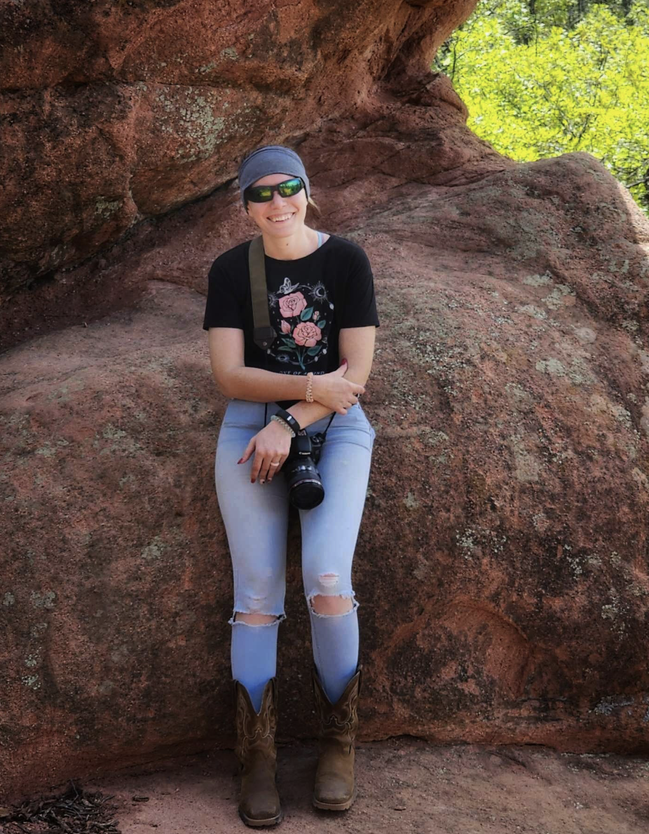 A woman sitting on a large red rock in a natural outdoor setting, wearing sunglasses, a hat, a black floral t-shirt, ripped jeans, and cowboy boots, smiling at the camera.