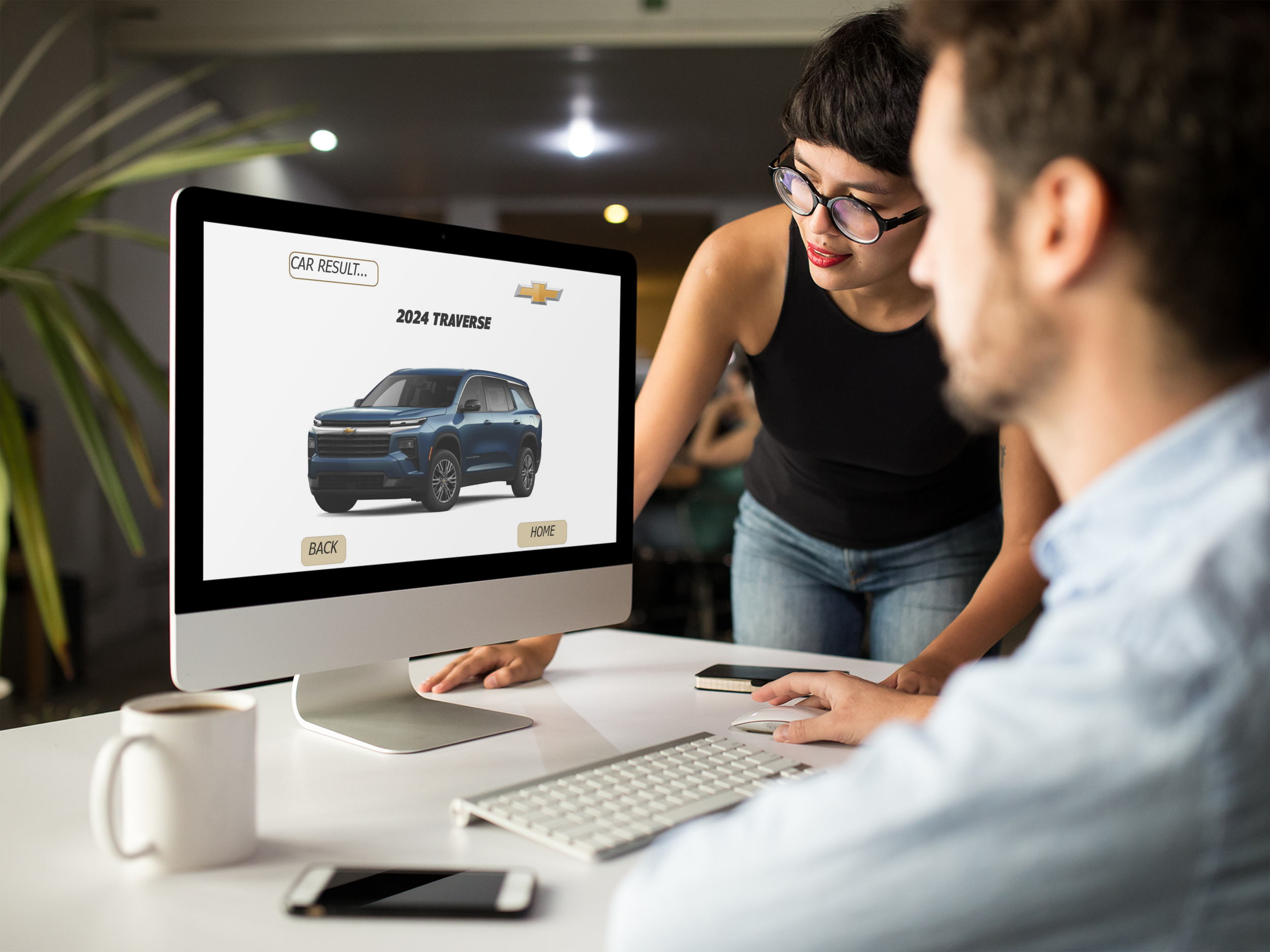 Two individuals reviewing a computer screen displaying a Chevrolet car model named "2024 Traverse." The woman with glasses and red lipstick looks at the screen while the man, partially visible, also focuses on it. The workspace has a white desk, a keyboard, a drink mug, and a smartphone.