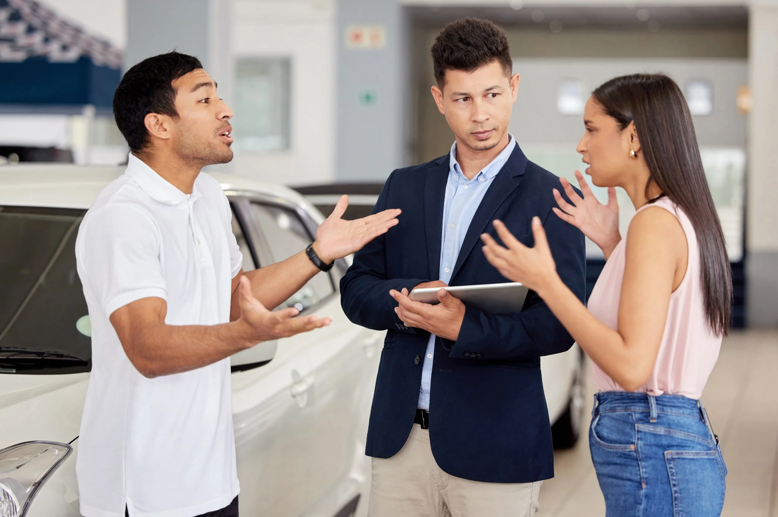 Three people having a conversation in a car dealership. One man appears to be explaining something, another man is holding a tablet, and a woman is gesturing with her hands.