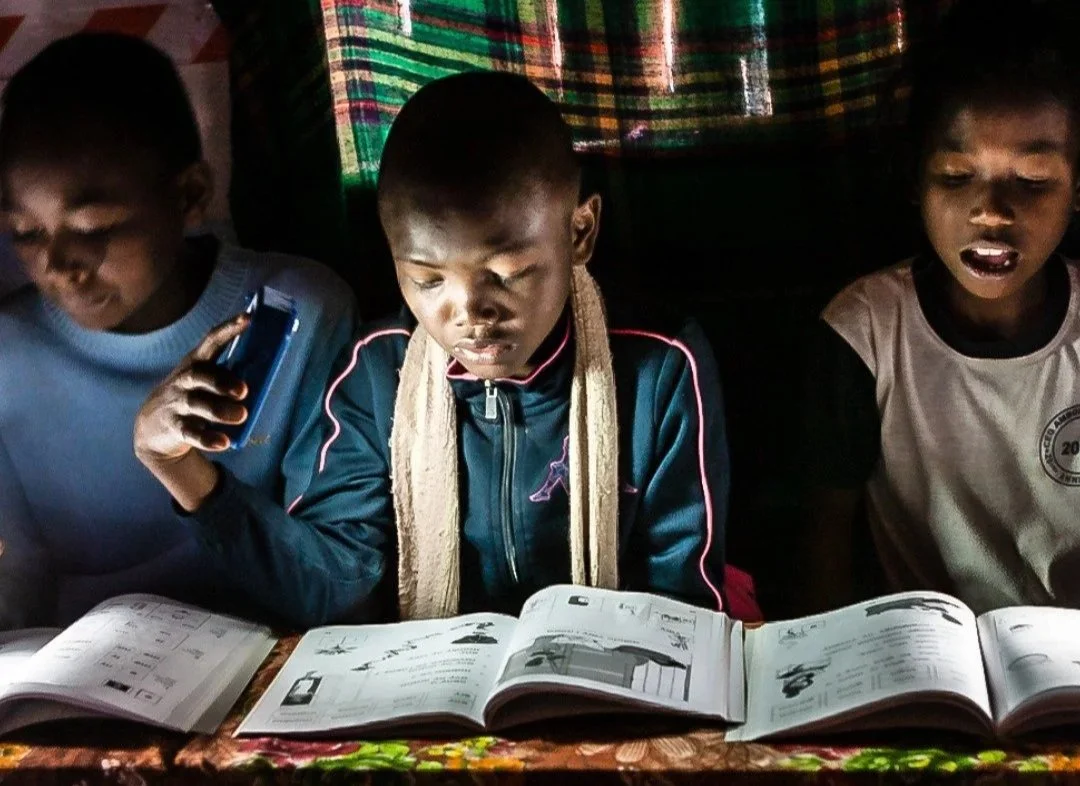 Three children reading from a textbook illuminated by a solar-powered lamp, demonstrating how portable renewable energy solutions enable education and learning opportunities in energy-poor communities while addressing energy poverty challenges.