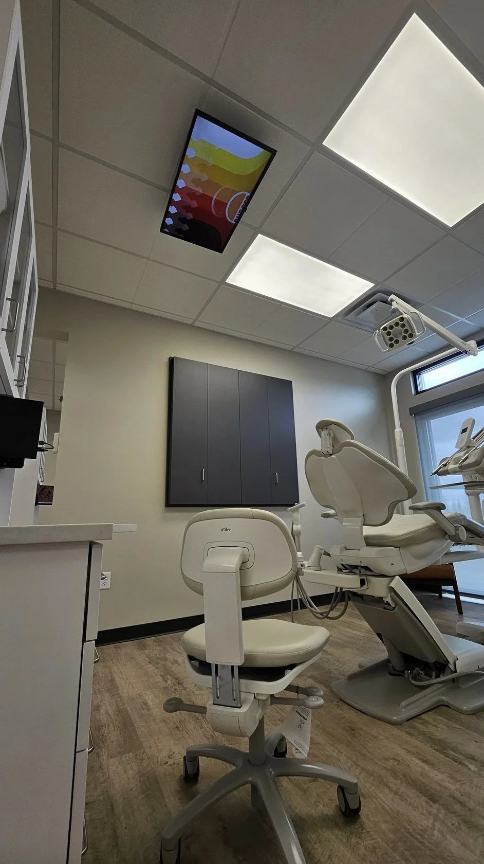 A dentist's office with a dental chair, a stool, a cabinet, and a wall-mounted storage unit. The ceiling has recessed lighting and a colorful digital display.