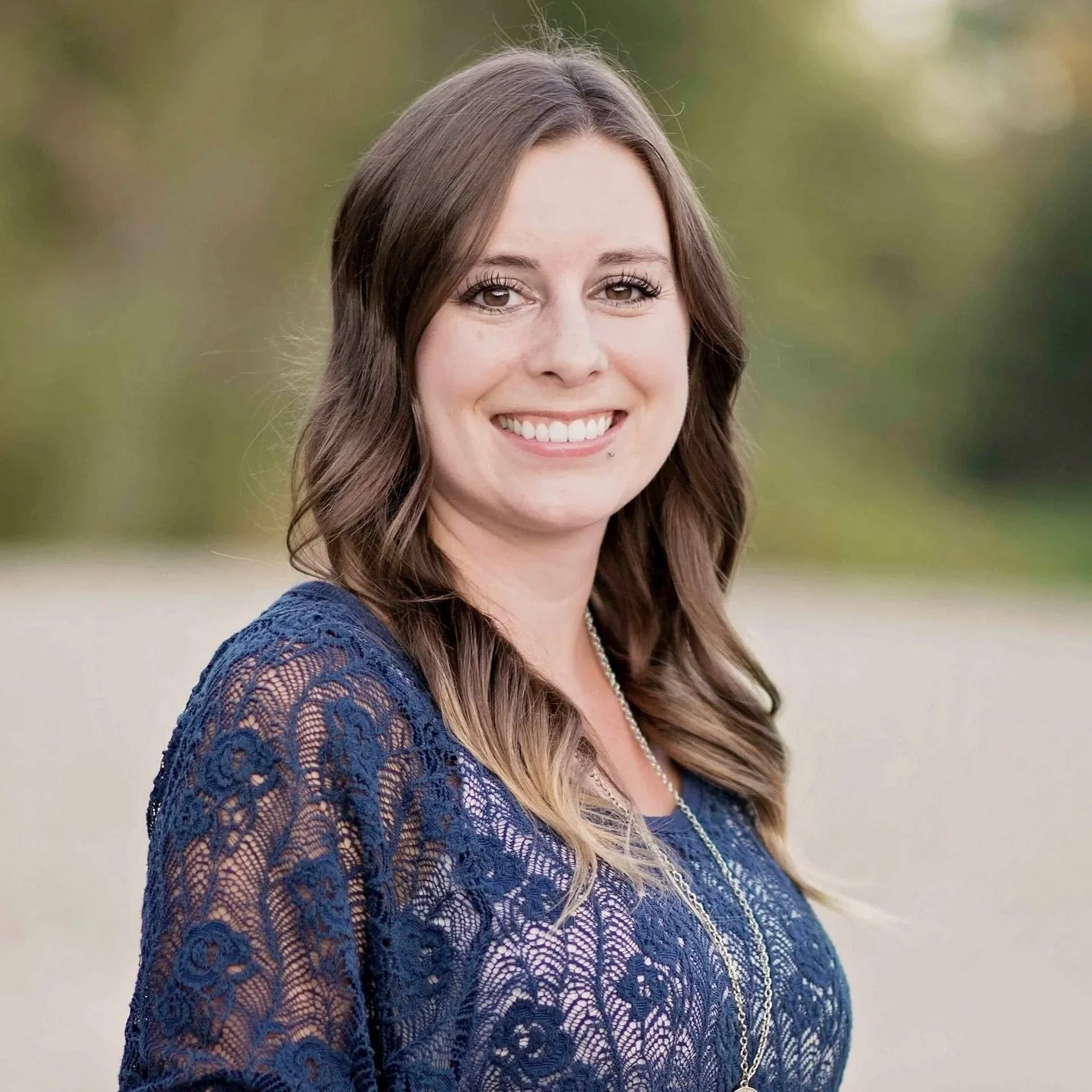 Portrait of a young woman with brown hair, smiling, wearing a blue lace dress and a pearl necklace, outdoors with a blurred background of green trees.