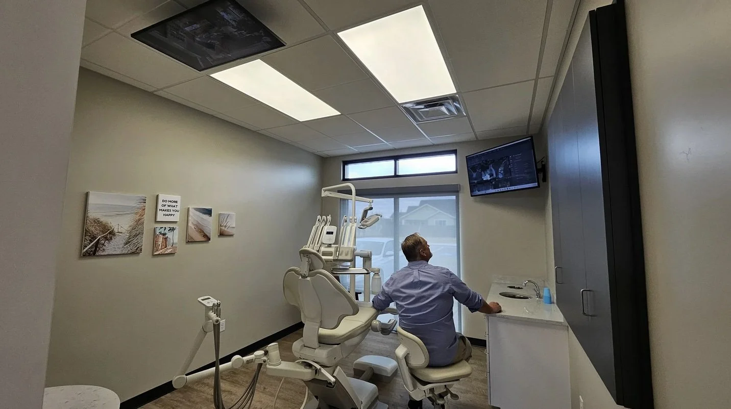 A man in a dental office sitting in a dental chair, looking at a monitor on the wall. The room has a window with blinds, some framed pictures and a sign on the wall that says 'Do more of what makes you happy.'