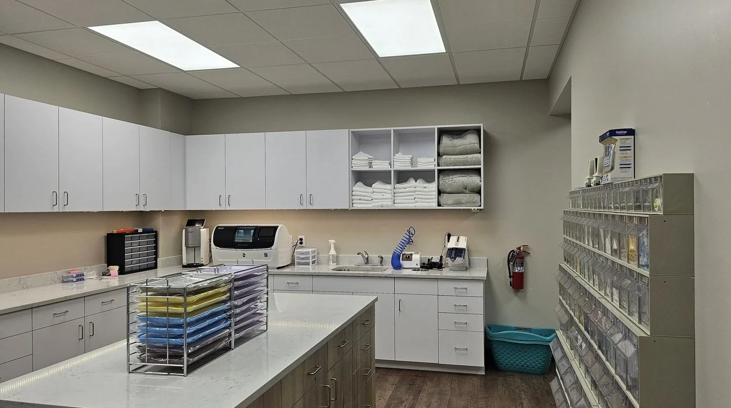 A medical supply room with white cabinets, shelves with folded towels and blankets, a countertop with a printer, disinfectant, and other supplies, a fire extinguisher, a laundry basket, and a wall-mounted organizer with small labeled compartments.
