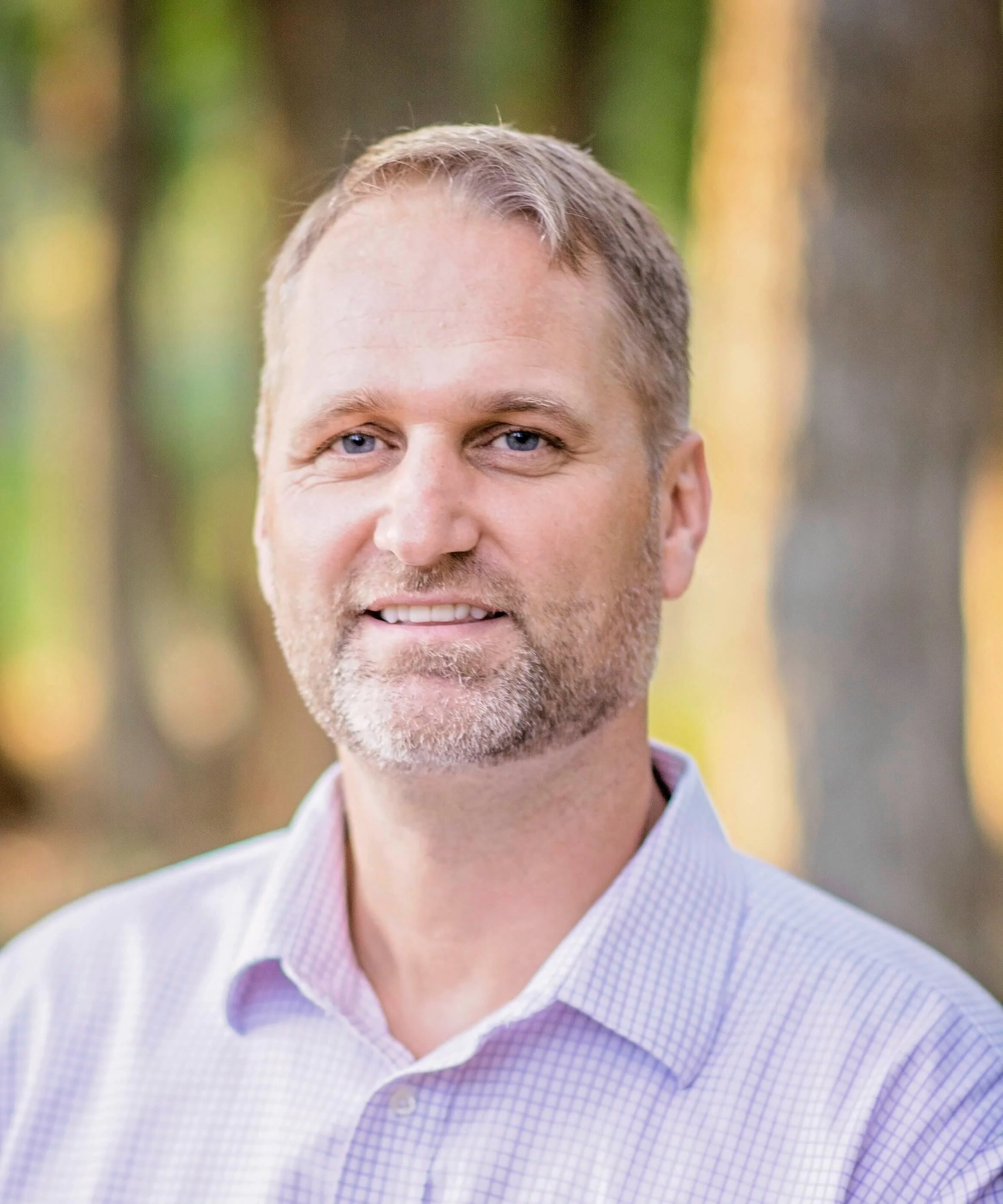 Smiling man with short hair and beard, wearing a light-colored shirt, outdoors with blurred greenery in the background.