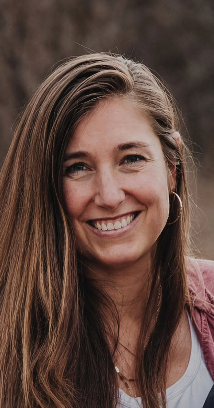 Close-up of a Katie Ripplinger, office manager at Whitehaven Dental. She has long brown hair smiling outdoors in natural light, wearing hoop earrings and a casual outfit.