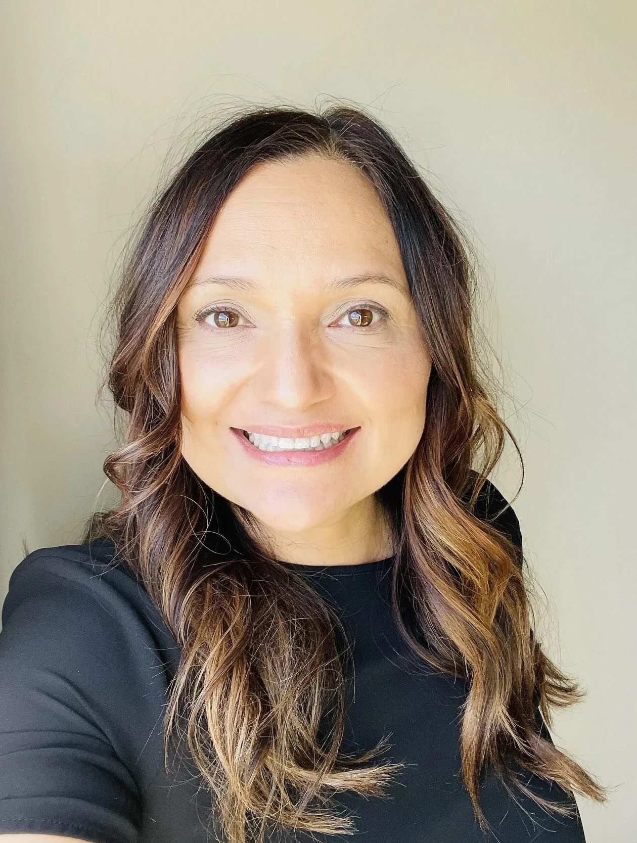 Lupe, a hygienist at Whitehaven Dental, with long brown hair and a big smile, wearing a black top, standing in front of a light-colored wall.
