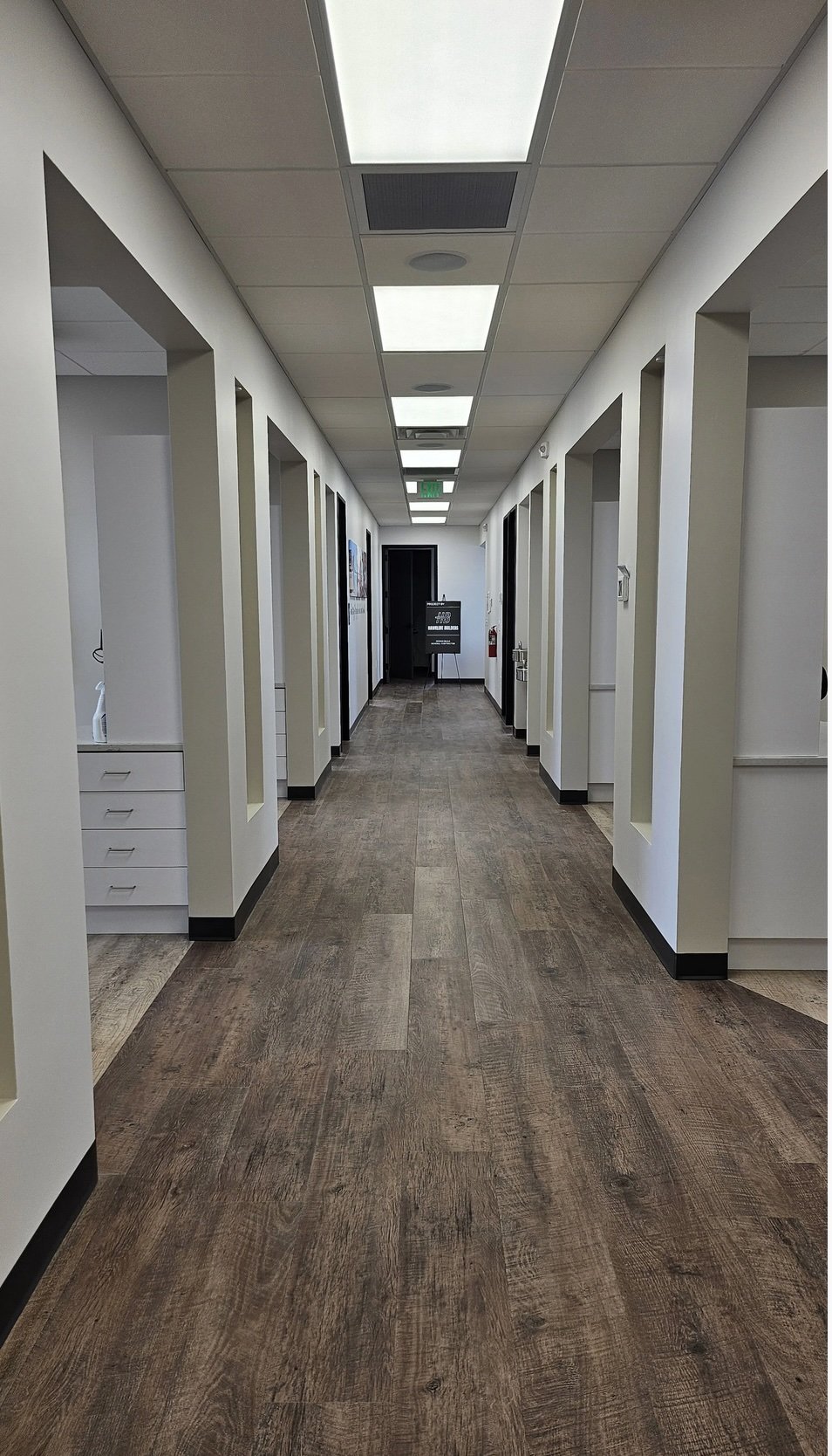 Empty hallway in an office or medical building with wood-like flooring, white walls, and ceiling lights.