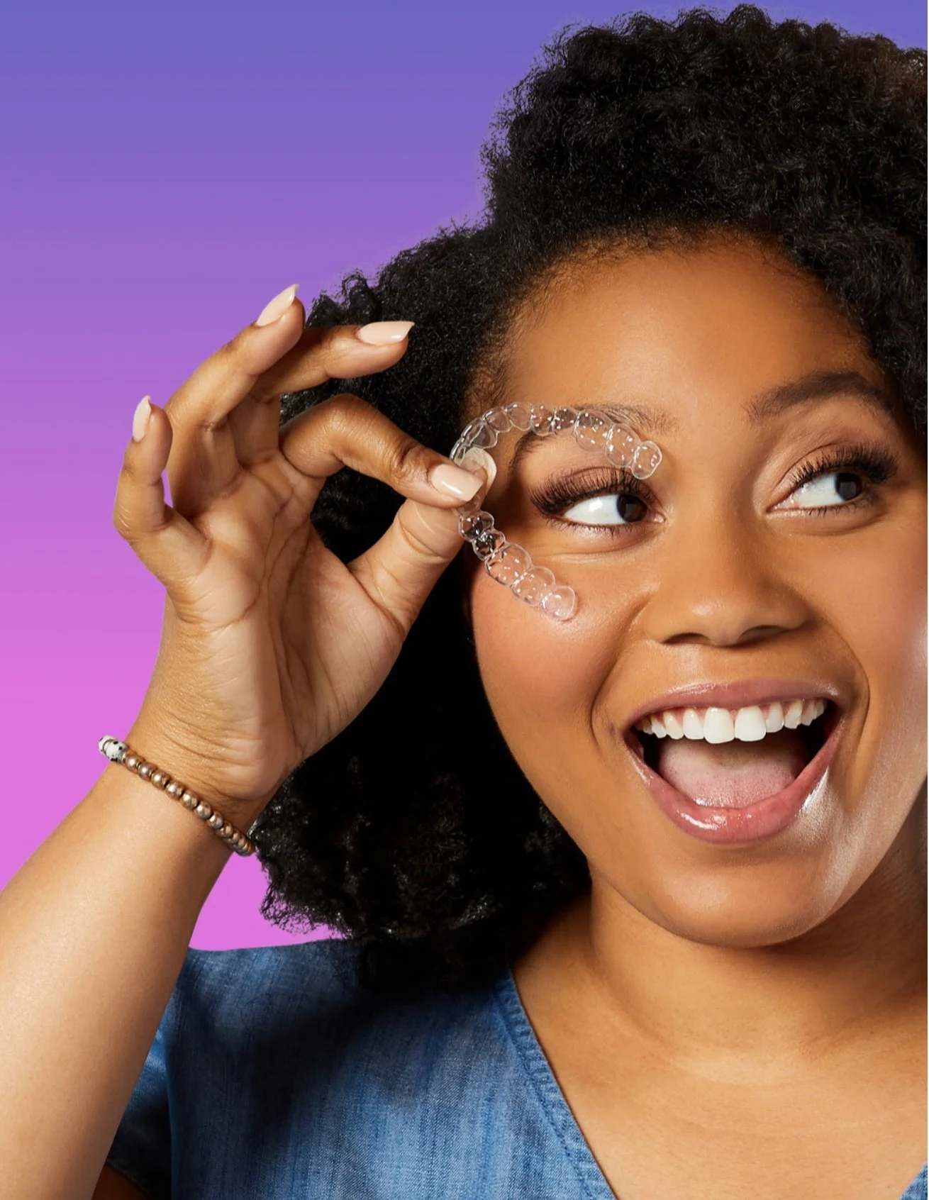 A woman with curly hair smiling and holding a clear, curved dental aligner near her eye.