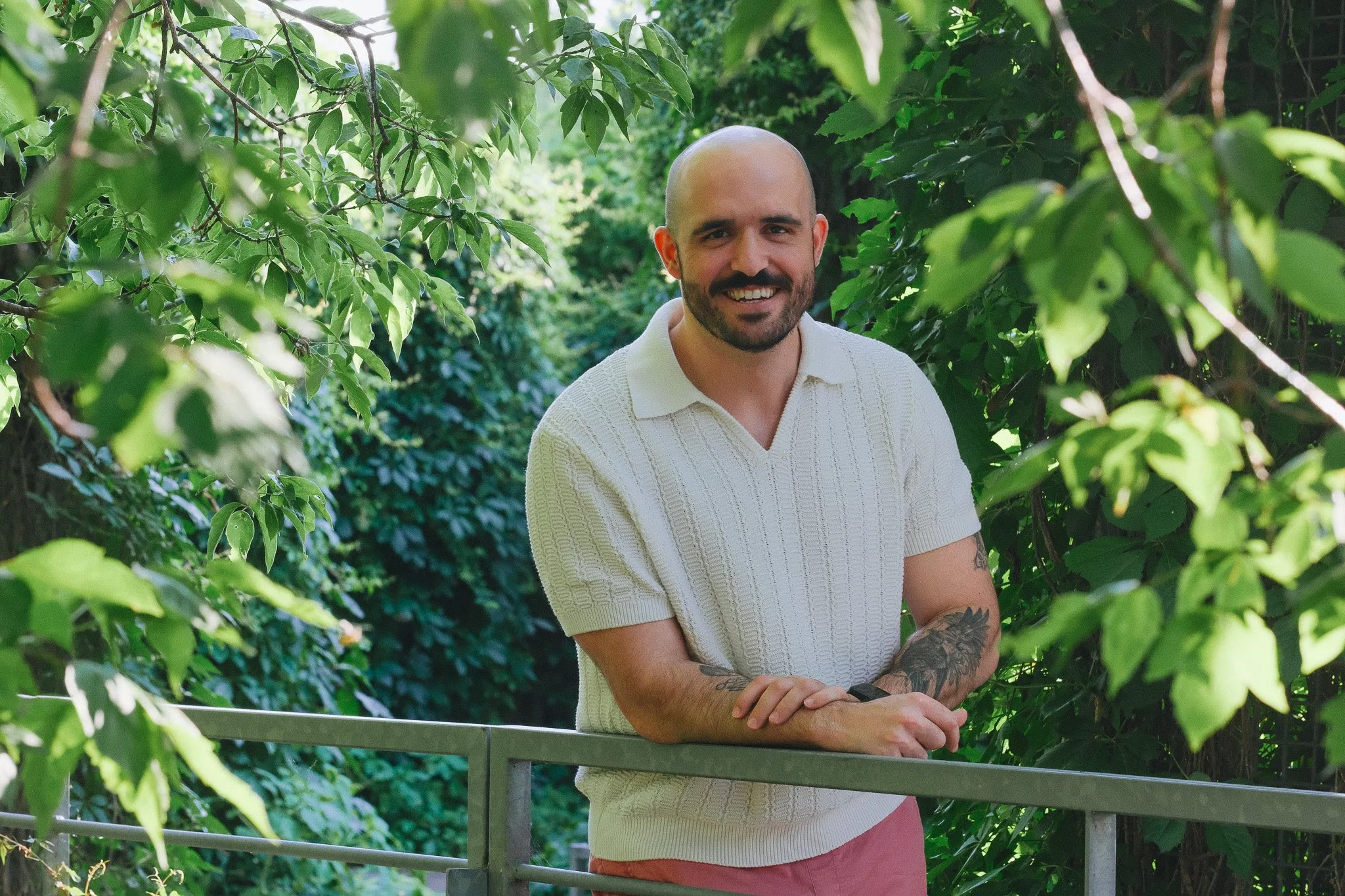 Un homme souriant avec une barbe, portant un pull blanc, en train de se tenir à une barrière dans un environnement verdoyant avec beaucoup de feuilles.