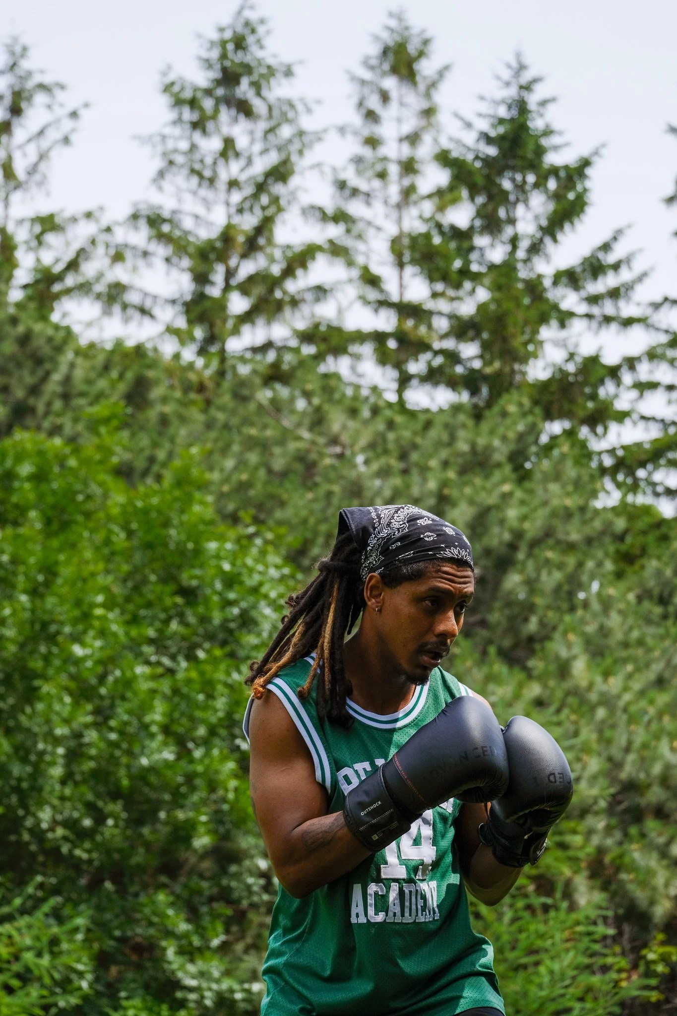 Un homme noir avec des dreadlocks portant un bandana noir s'entraînant à la boxe avec des gants de boxe noirs dans un environnement naturel avec des arbres en arrière-plan.