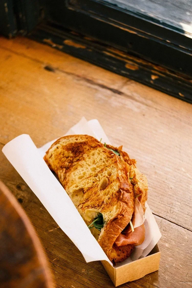 Sandwich baguette avec jambon, fromage et légumes dans une boîte en carton, sur une table en bois.