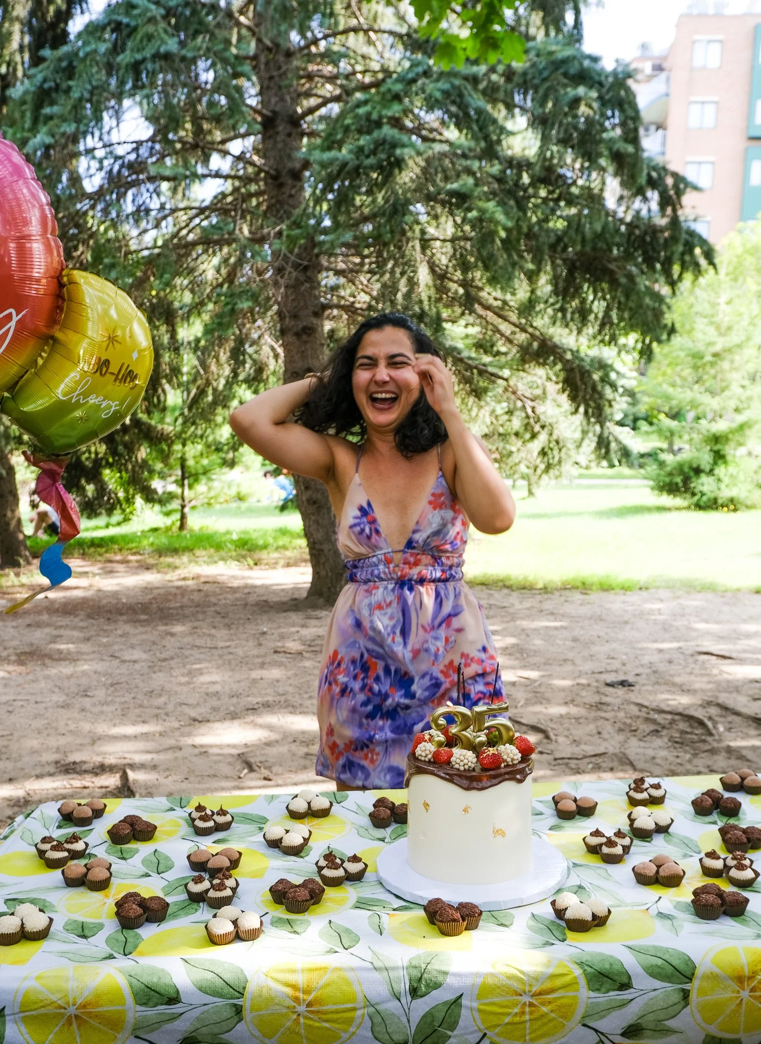 Une femme souriante fête son anniversaire avec un gâteau et des cupcakes, devant un décor ambiant en plein air avec des ballons et une table ornée d'un nappage à motif agrumes.