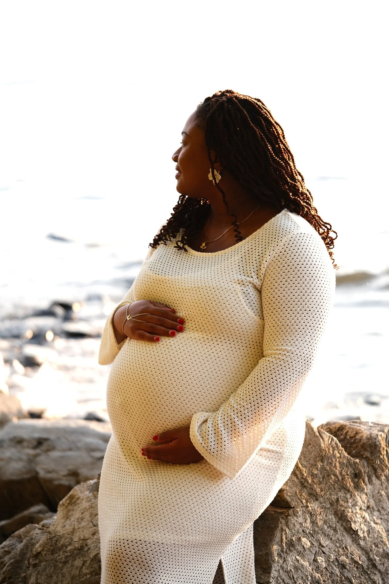 Une femme enceinte souriante se tient sur une plage rocheuse au coucher du soleil, portant une robe blanche en maille et des bijoux.