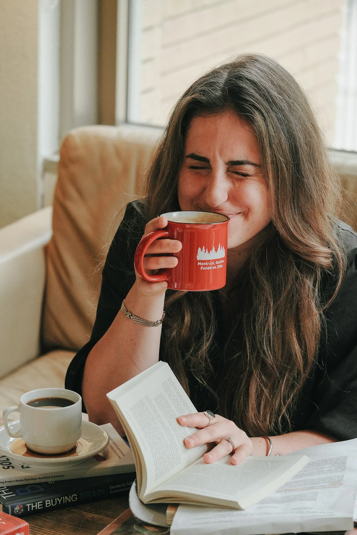 Une jeune femme avec de longs cheveux bruns qui lit un livre ouvert tout en buvant une tasse de café ou de thé dans un café ou une salle de lecture.