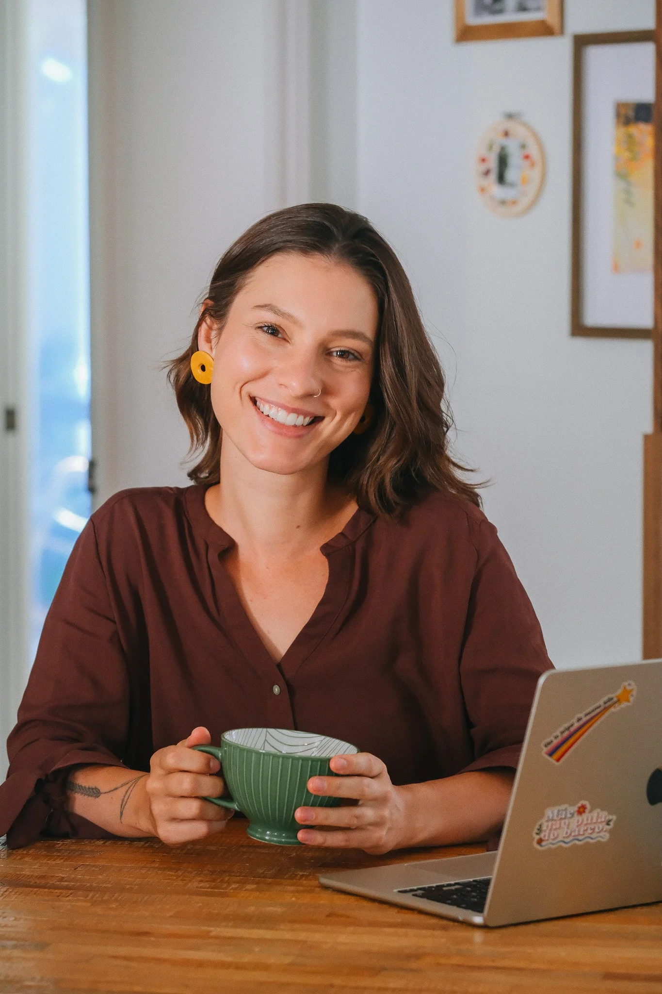 Une femme souriante tenant une tasse verte, assise à une table en bois, avec un ordinateur portable à ses côtés dans une pièce lumineuse.