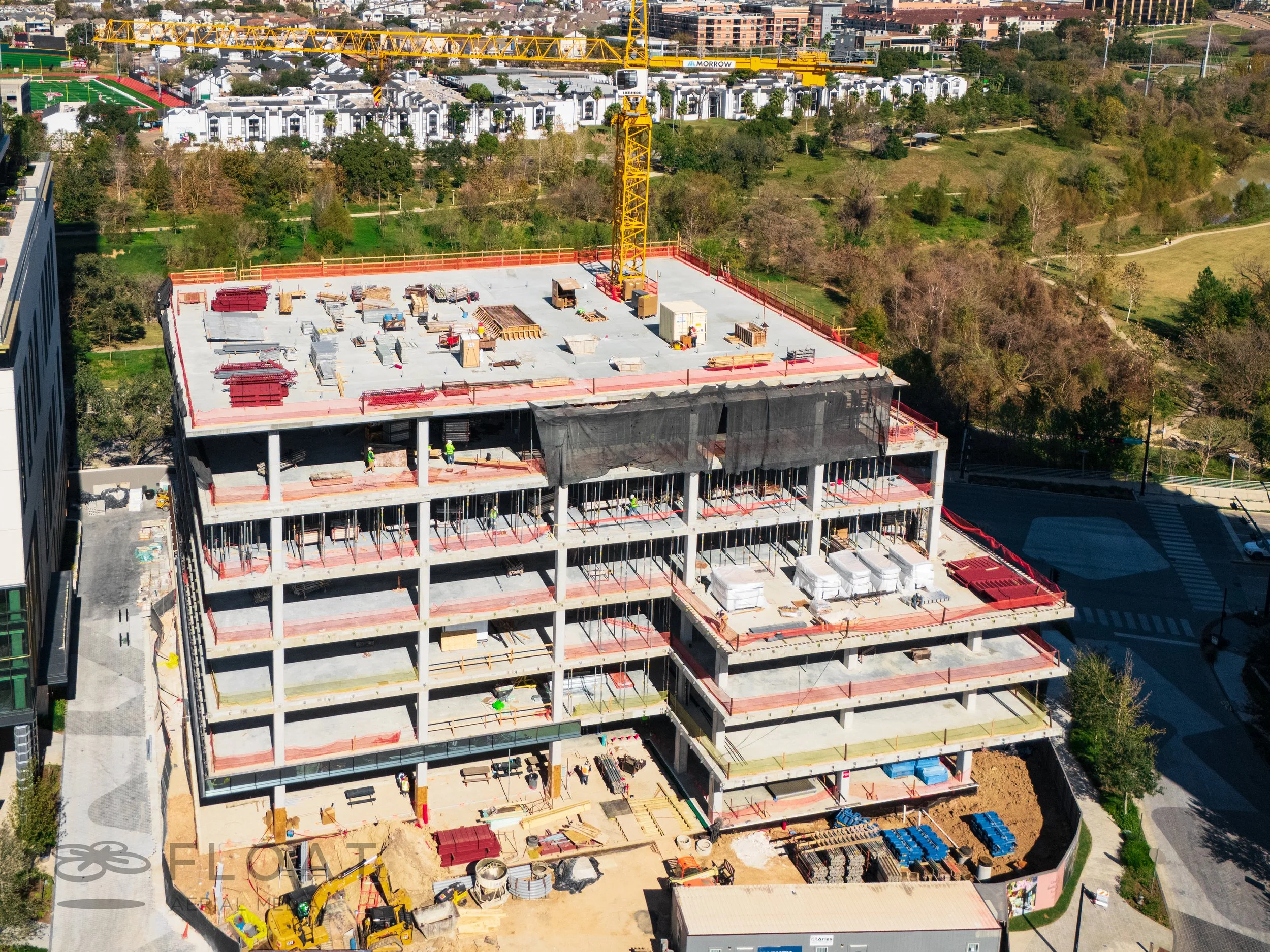 A multi-story building under construction with a yellow crane on top, surrounded by trees and city buildings.