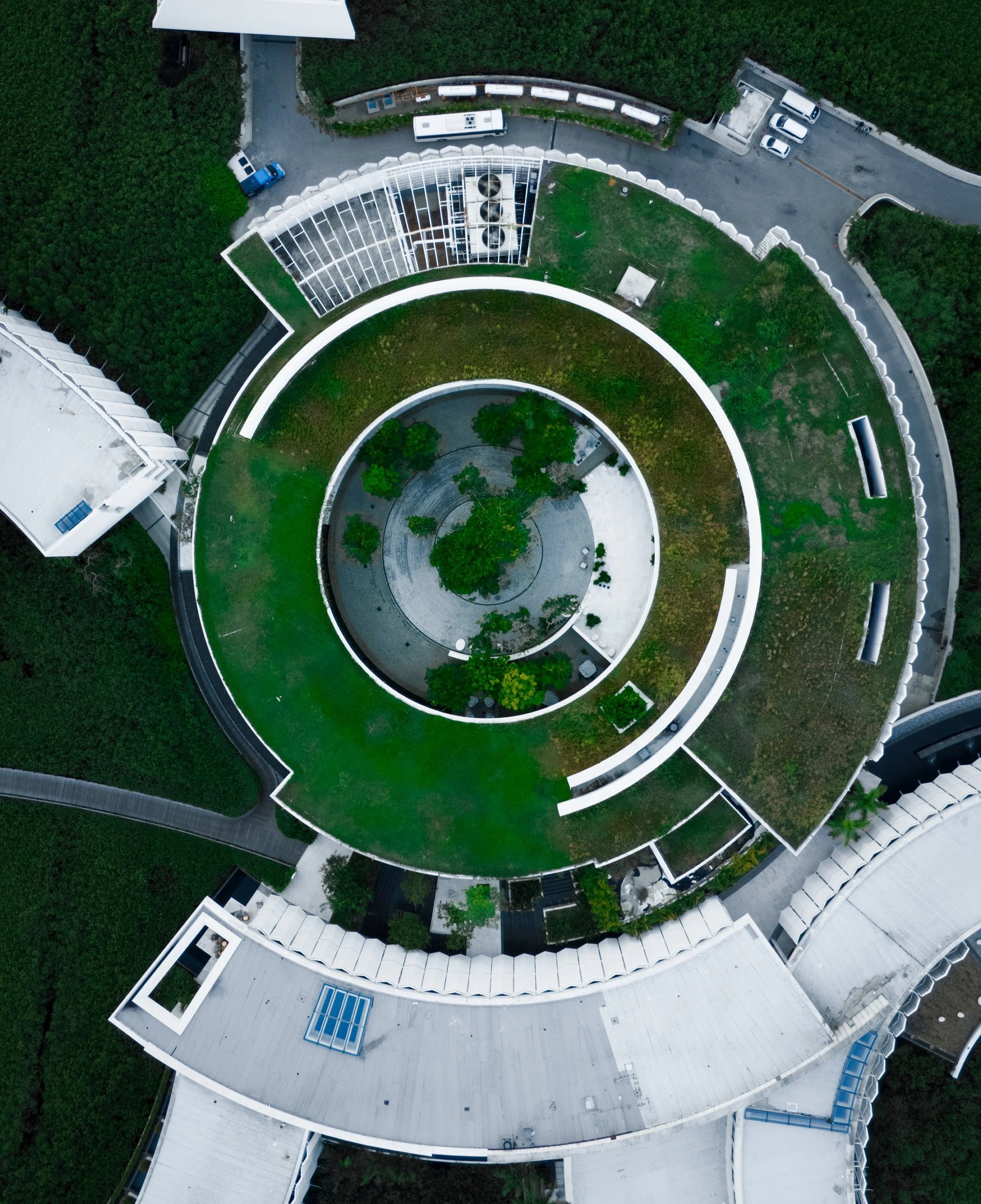 Aerial view of a modern, circular building with green rooftop gardens and trees at the center, surrounded by white and gray structures and parking areas.