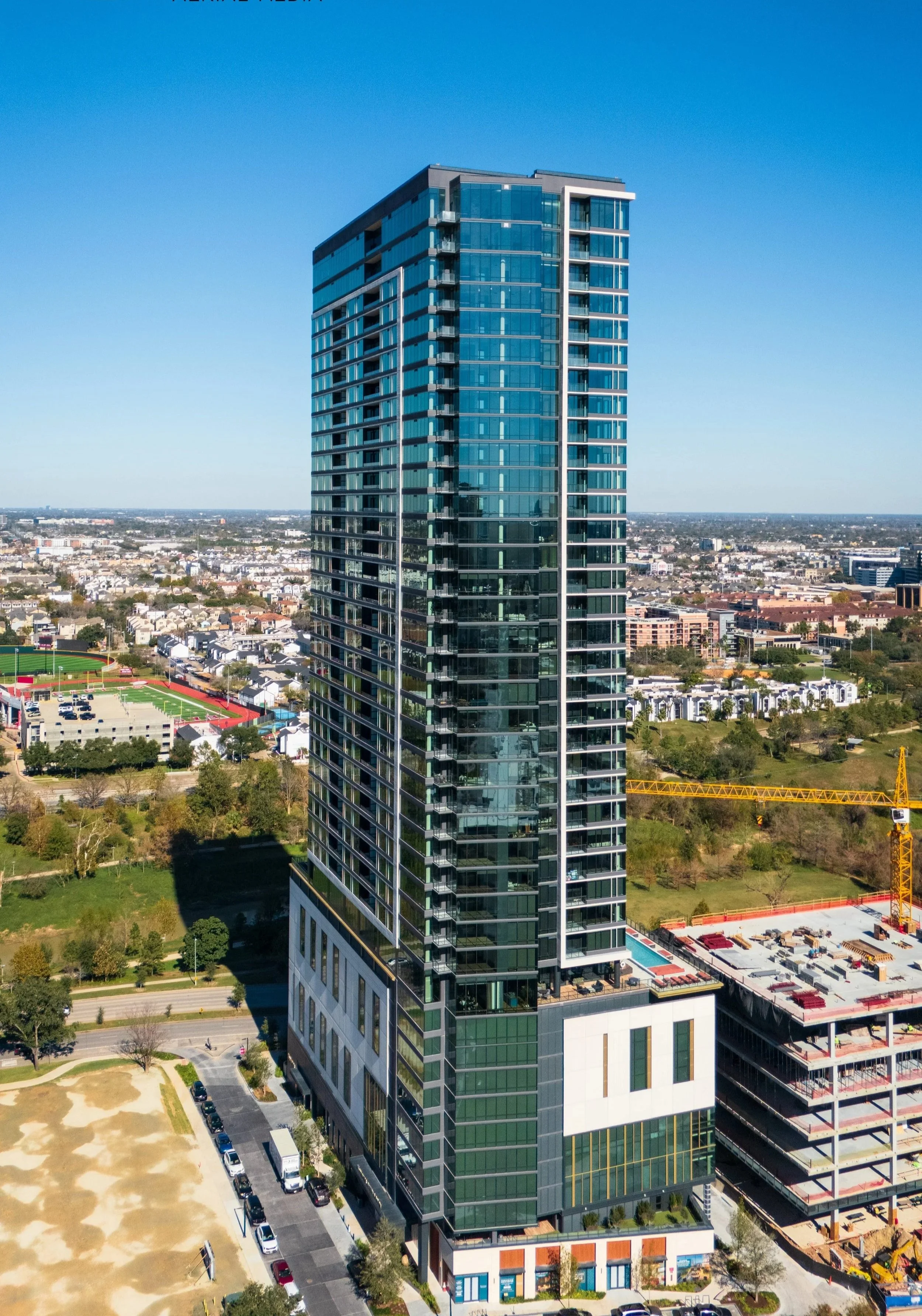 A tall modern glass skyscraper in an urban area with parks, a construction site, and other buildings in the background under a clear blue sky.