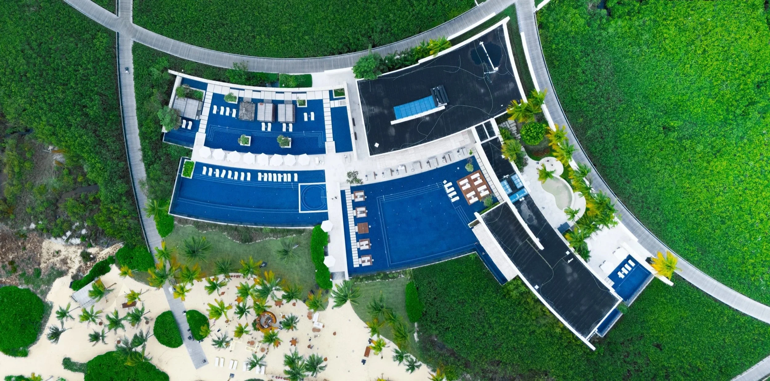 Aerial view of a modern resort with multiple swimming pools, a basketball court, surrounded by green landscape and palm trees.