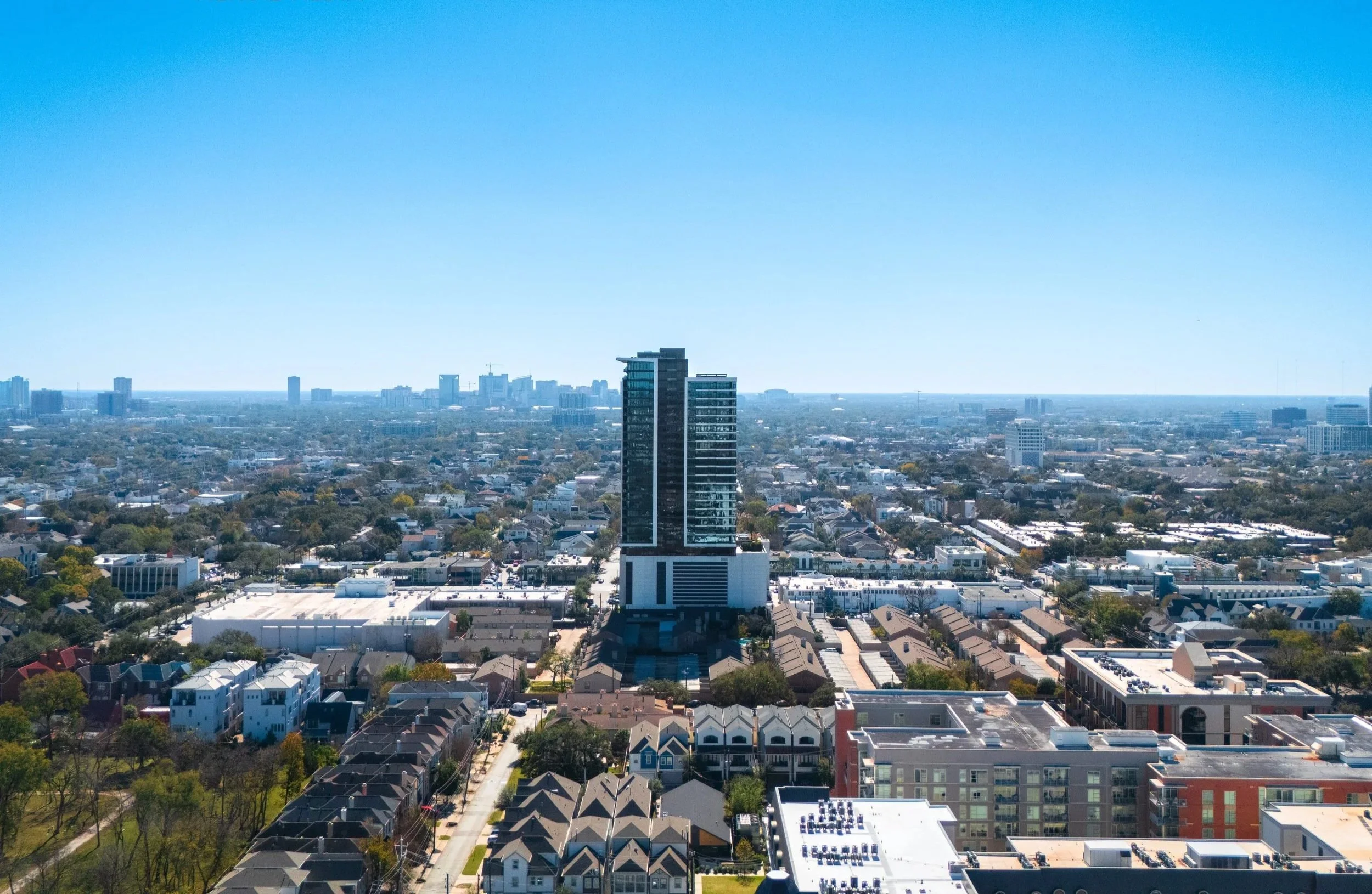 Aerial view of a city with a tall modern building in the center, surrounded by smaller buildings, with a clear blue sky overhead.