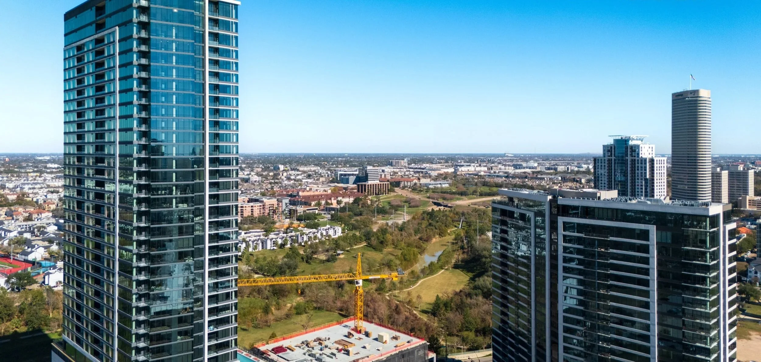Skyline view of downtown with tall modern glass skyscrapers, a yellow construction crane, parks, residential areas, and a clear blue sky.