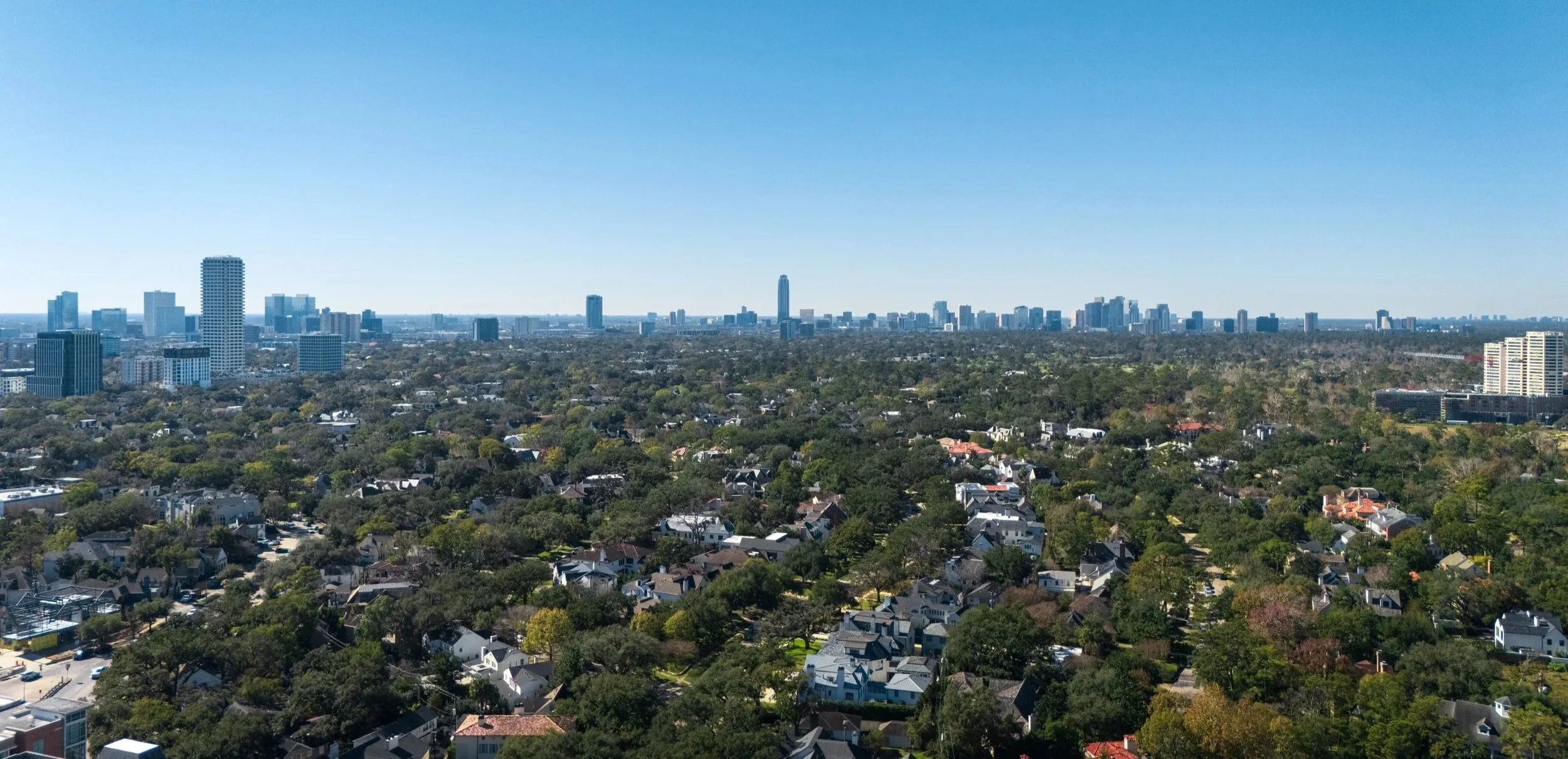 Aerial view of a city skyline with tall buildings in the distance and a suburban neighborhood with houses and trees in the foreground under a clear blue sky.