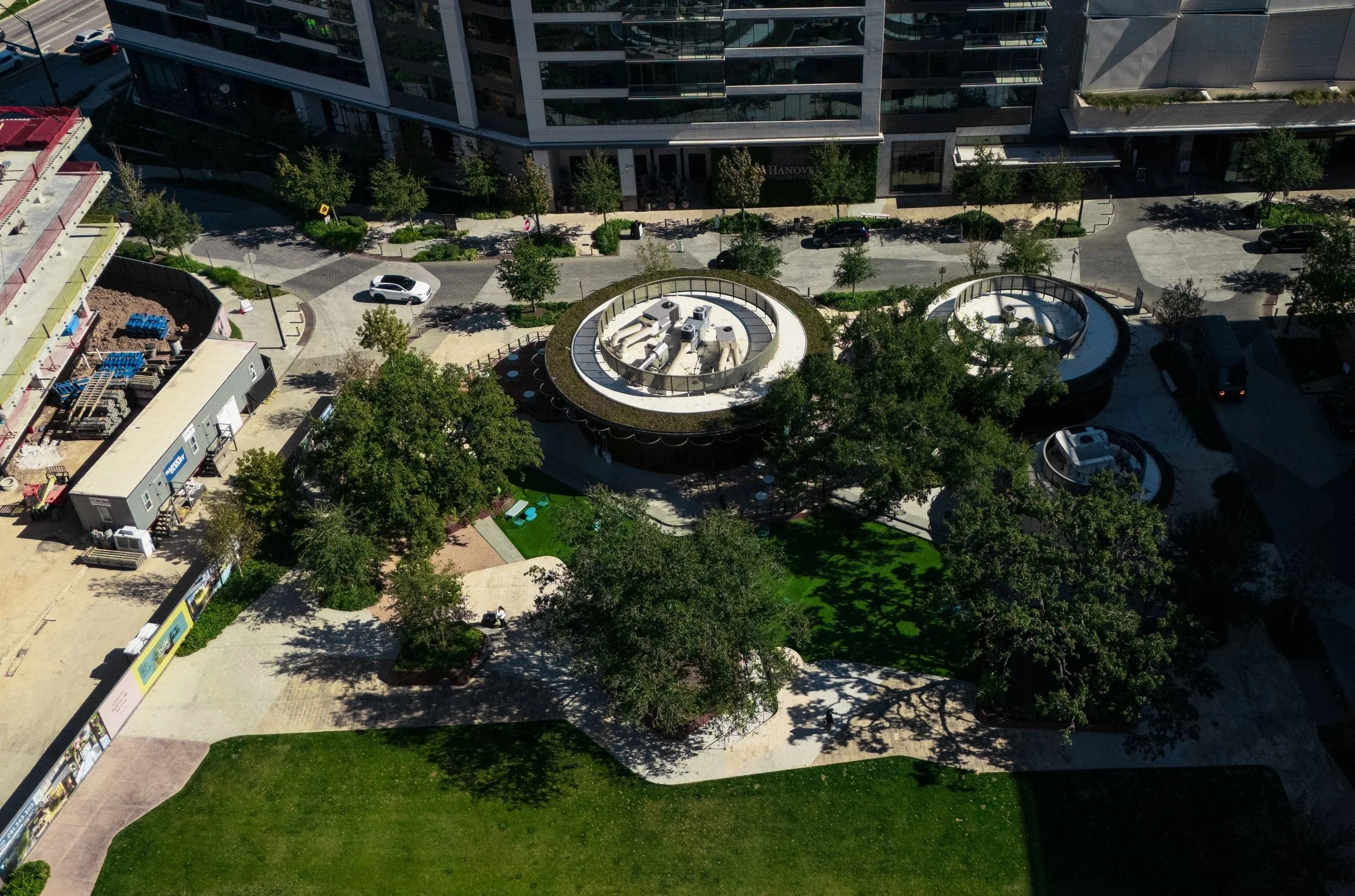 Aerial view of modern urban park with trees, walkways, and round architectural structures surrounded by office buildings, parking lots, and construction site.