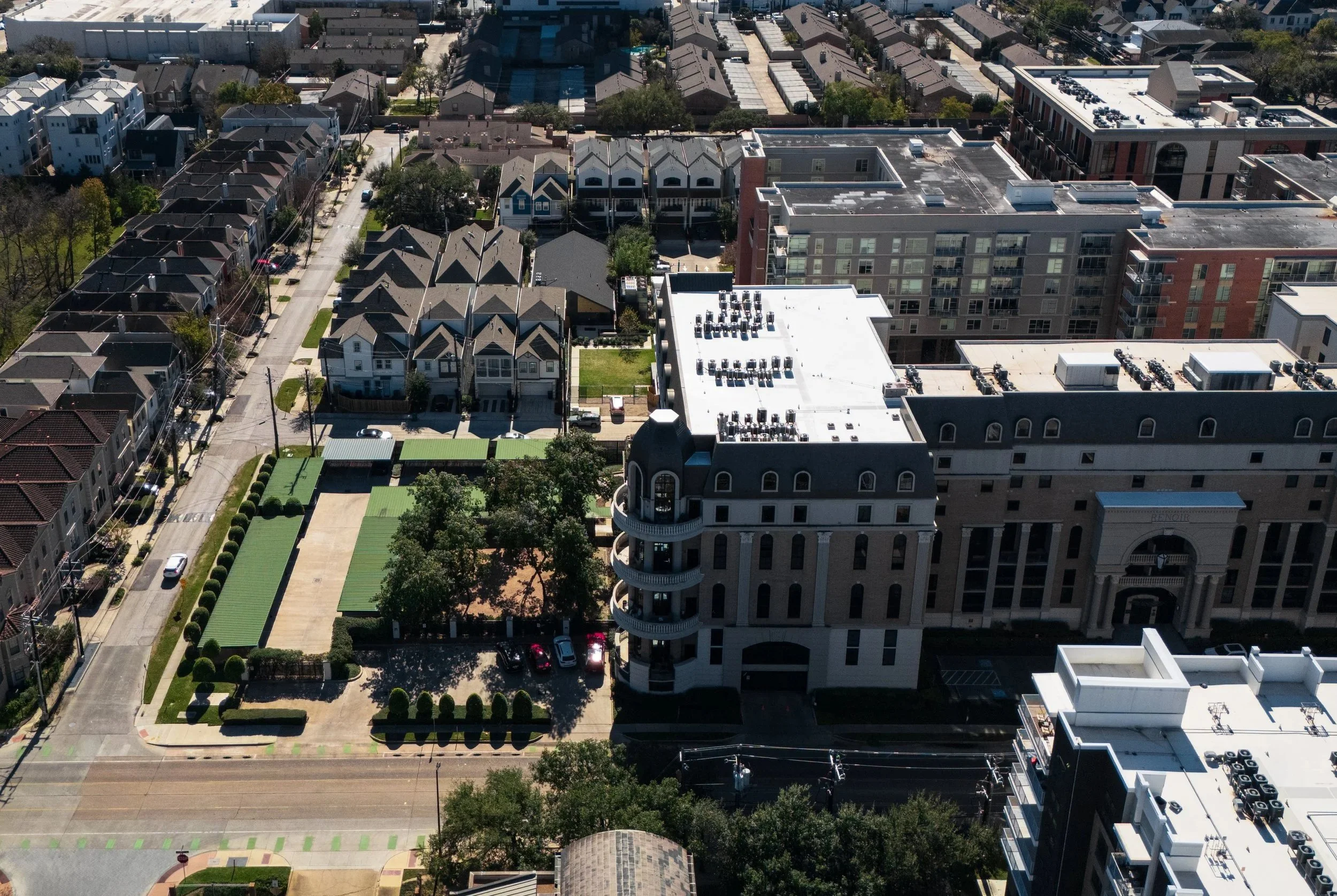 An aerial view of a city block showing a mix of residential houses and larger commercial or apartment buildings, with streets, parking lots, and greenery.