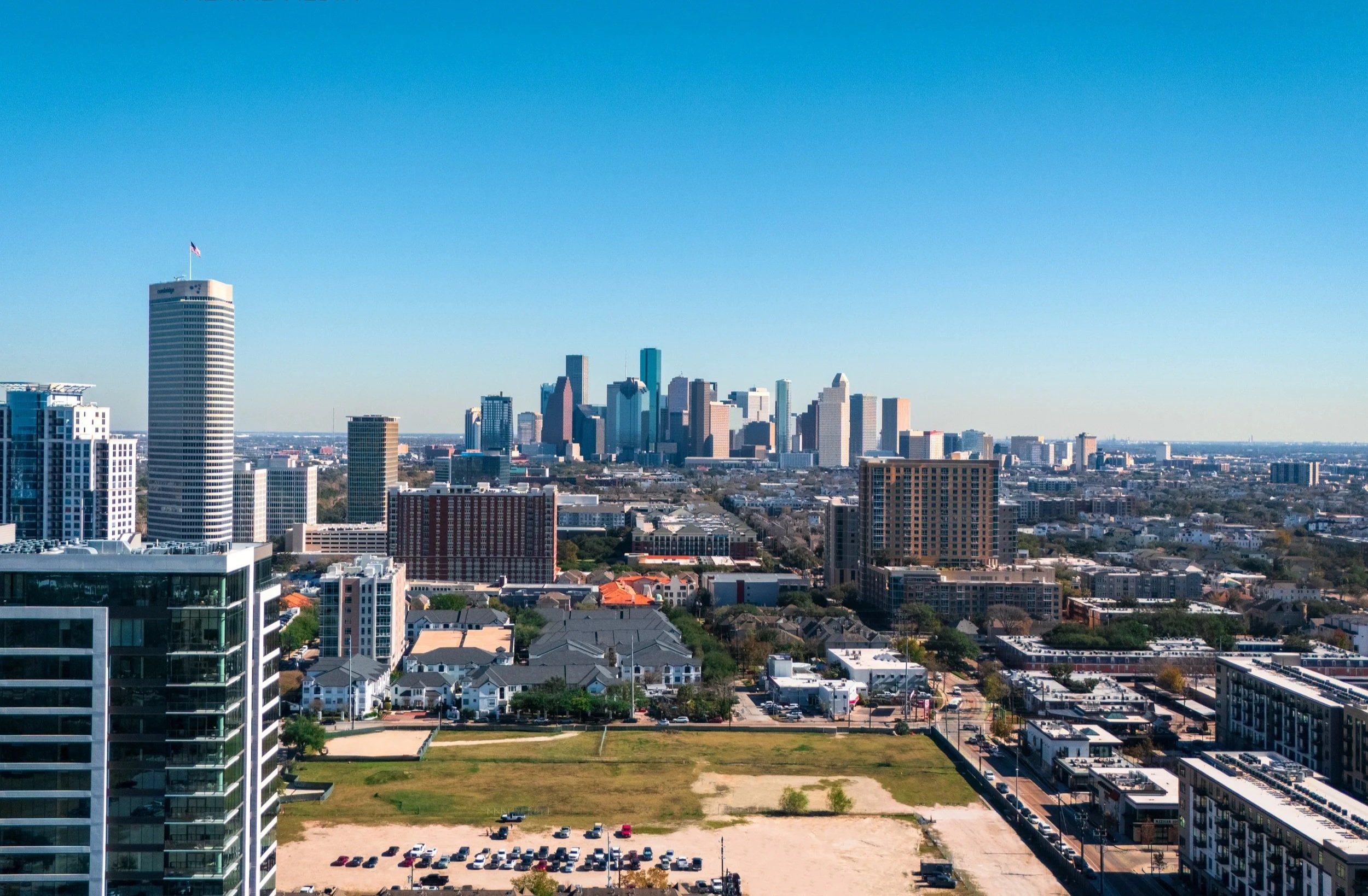 City skyline with tall skyscrapers and a clear blue sky, with a mix of modern buildings, residential houses, and green spaces in the foreground.
