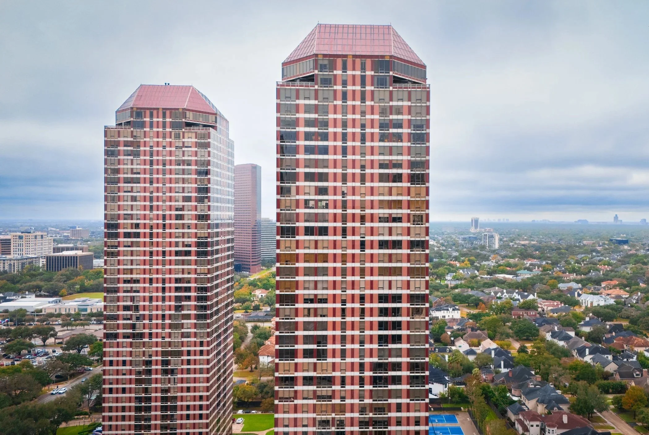 Two tall, modern, red and gray skyscrapers with cropping windows, overlooking a suburban neighborhood with houses and trees, and a city skyline in the distance under cloudy skies.